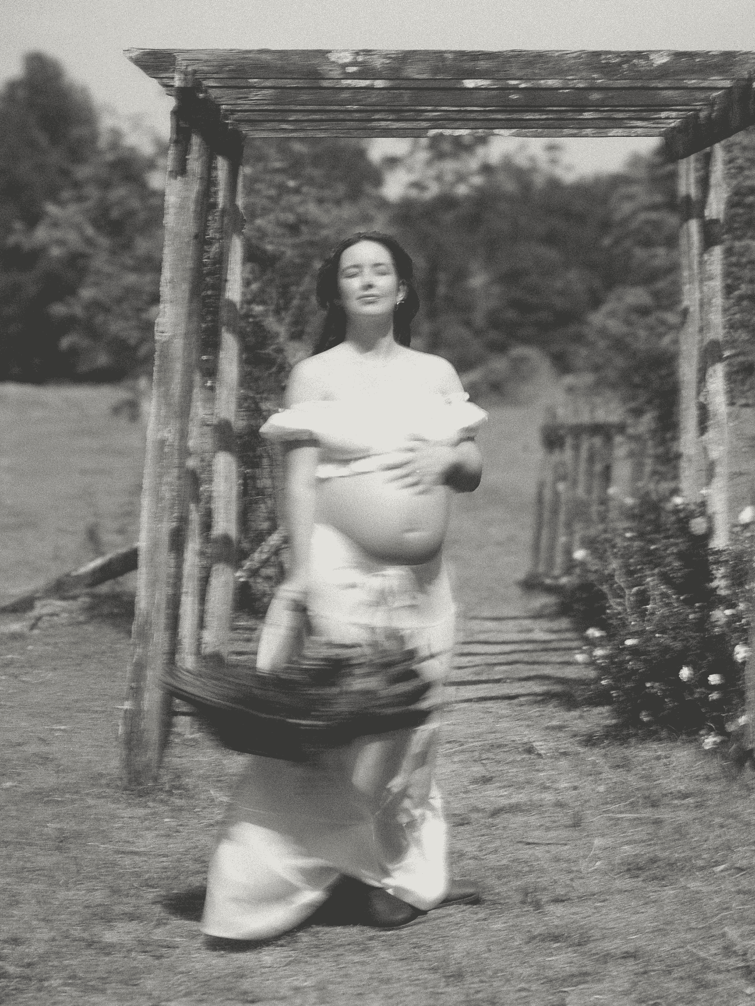 Black-and-white photo of a woman kneeling on the grass under a wooden structure with trees in the background.