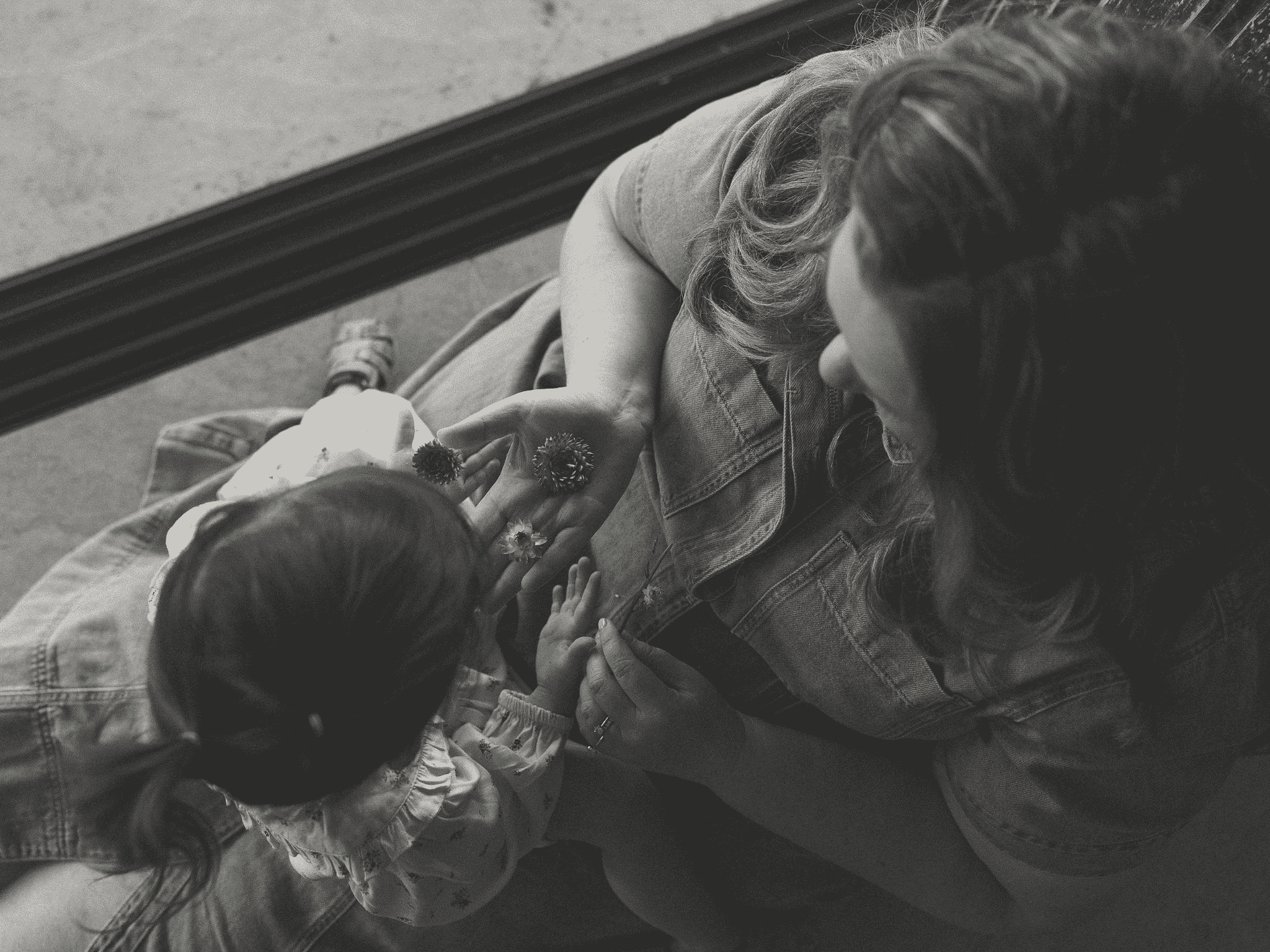 A black-and-white photograph of two children, one boy and one girl, sitting on the ground. The girl is leaning over the boy, and they are holding hands with a flower between them.