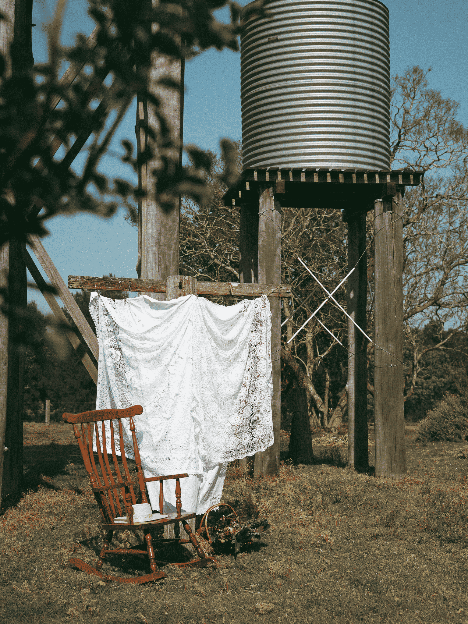 An outdoor scene with a wooden chair, a white lace cloth hanging from a string, a pair of shoes, and an electric water tower in the background.