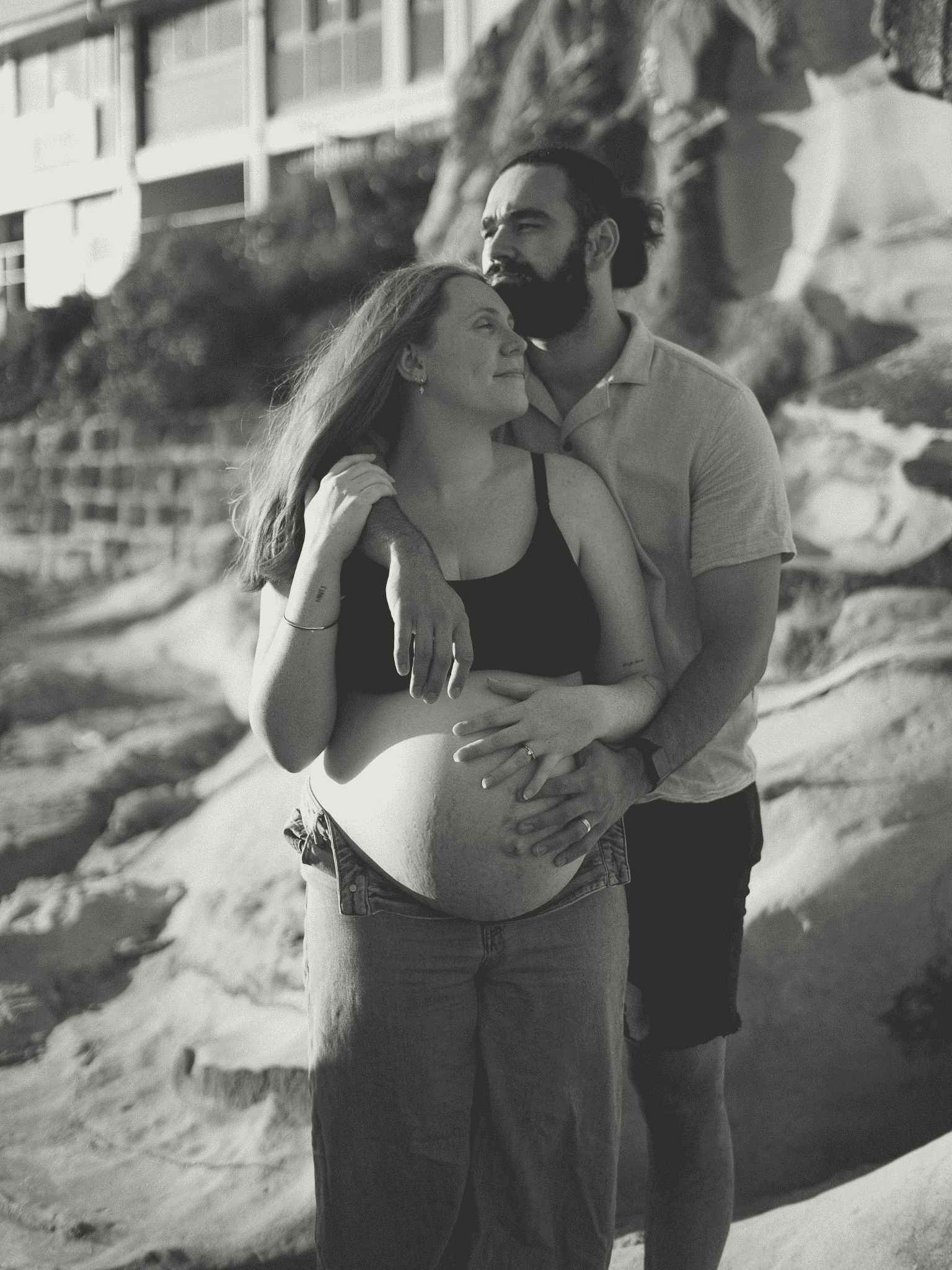 A couple standing on a beach with rocks and a building in the background. The woman, with long hair, is pregnant and is being embraced by the man. The woman is smiling with eyes closed, and the man is touching her head and smiling with eyes closed.