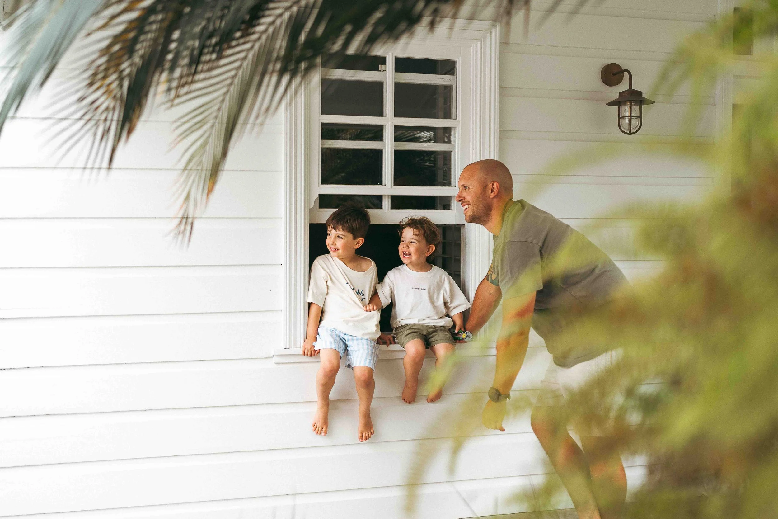 Two young boys looking out of a window with toy animals, a rabbit and a dinosaur, positioned on the window sill. The boys are dressed in white T-shirts and appear curious or playful.
