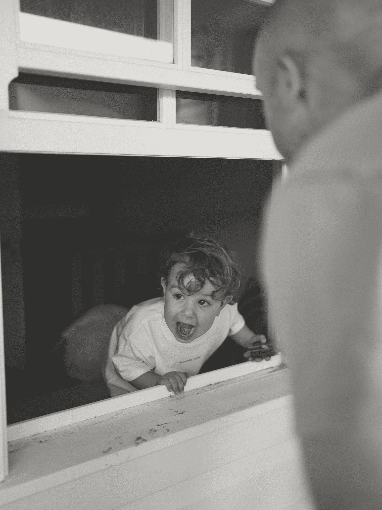 A young boy with curly hair is looking out a window with an excited expression, mouth open in a smile or shout, leaning on the window sill with one hand.