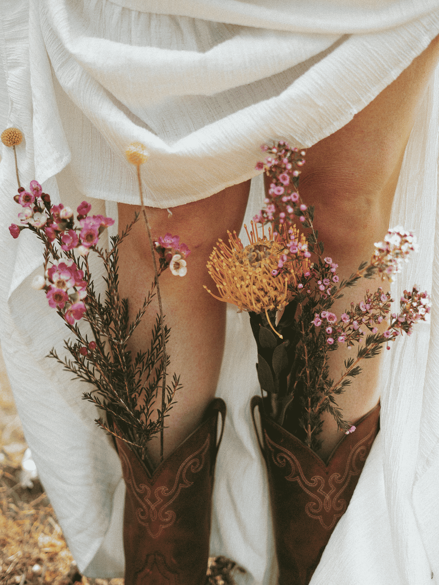 Close-up of a person wearing a white garment with embroidered details, holding a bouquet of pink and yellow flowers in front of their legs, which are clad in brown boots with decorative stitching.