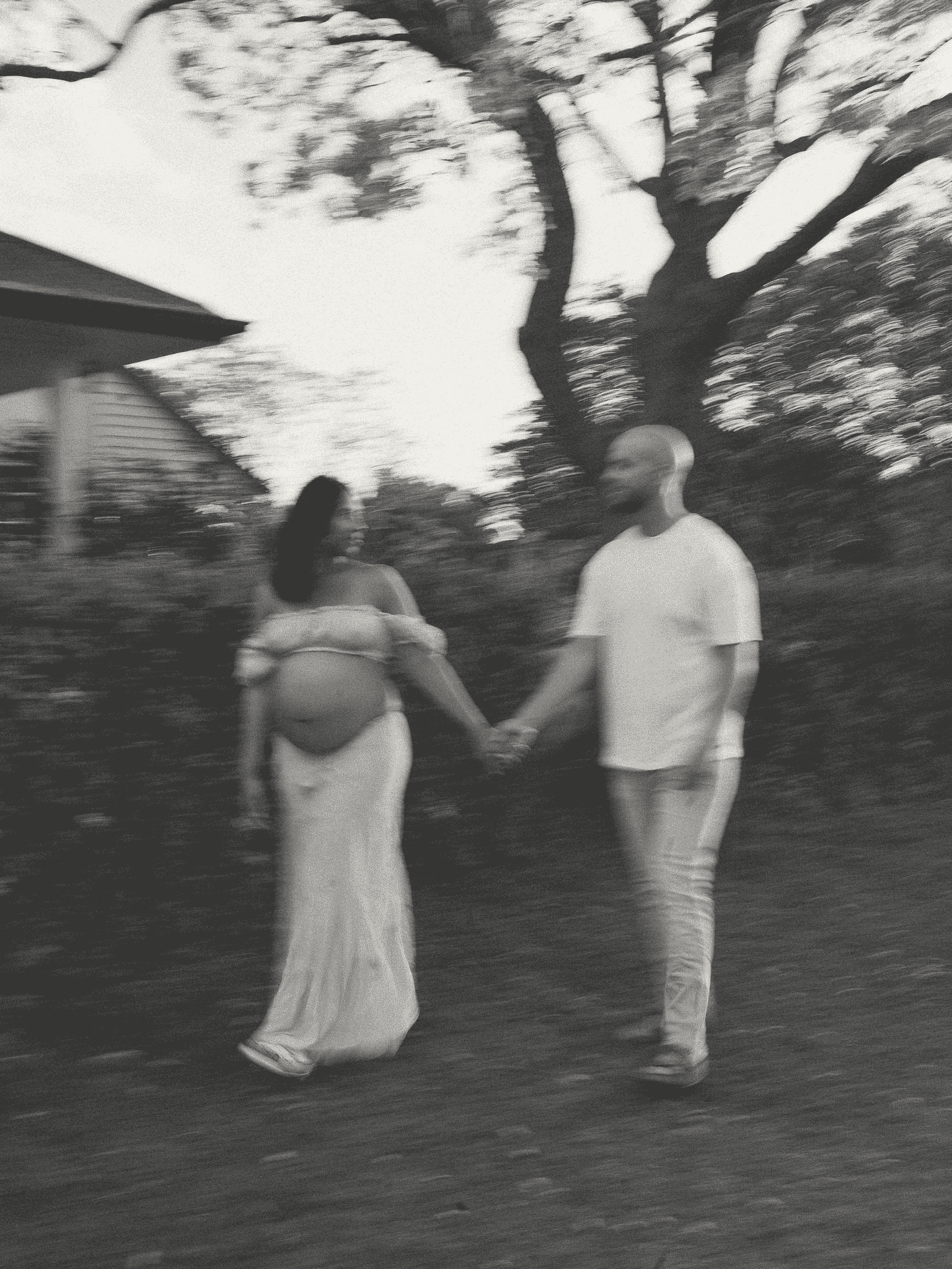 A couple holding hands and walking on a grassy outdoor area with a large tree in the background. The woman is pregnant and wearing a long skirt and off-the-shoulder top. The man is dressed in a t-shirt and jeans.