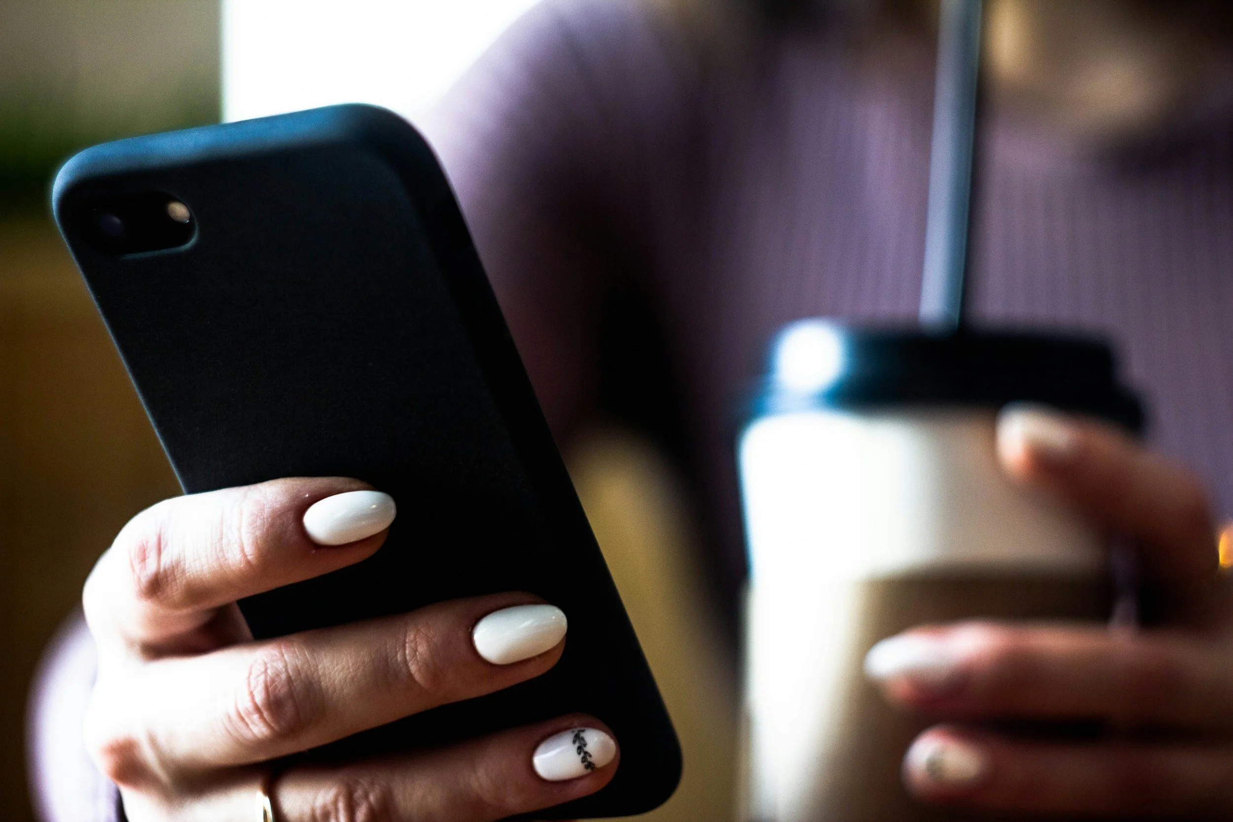 A person holding a smartphone with a black case. The person has white painted nails with a small decorative design. The background is blurry.