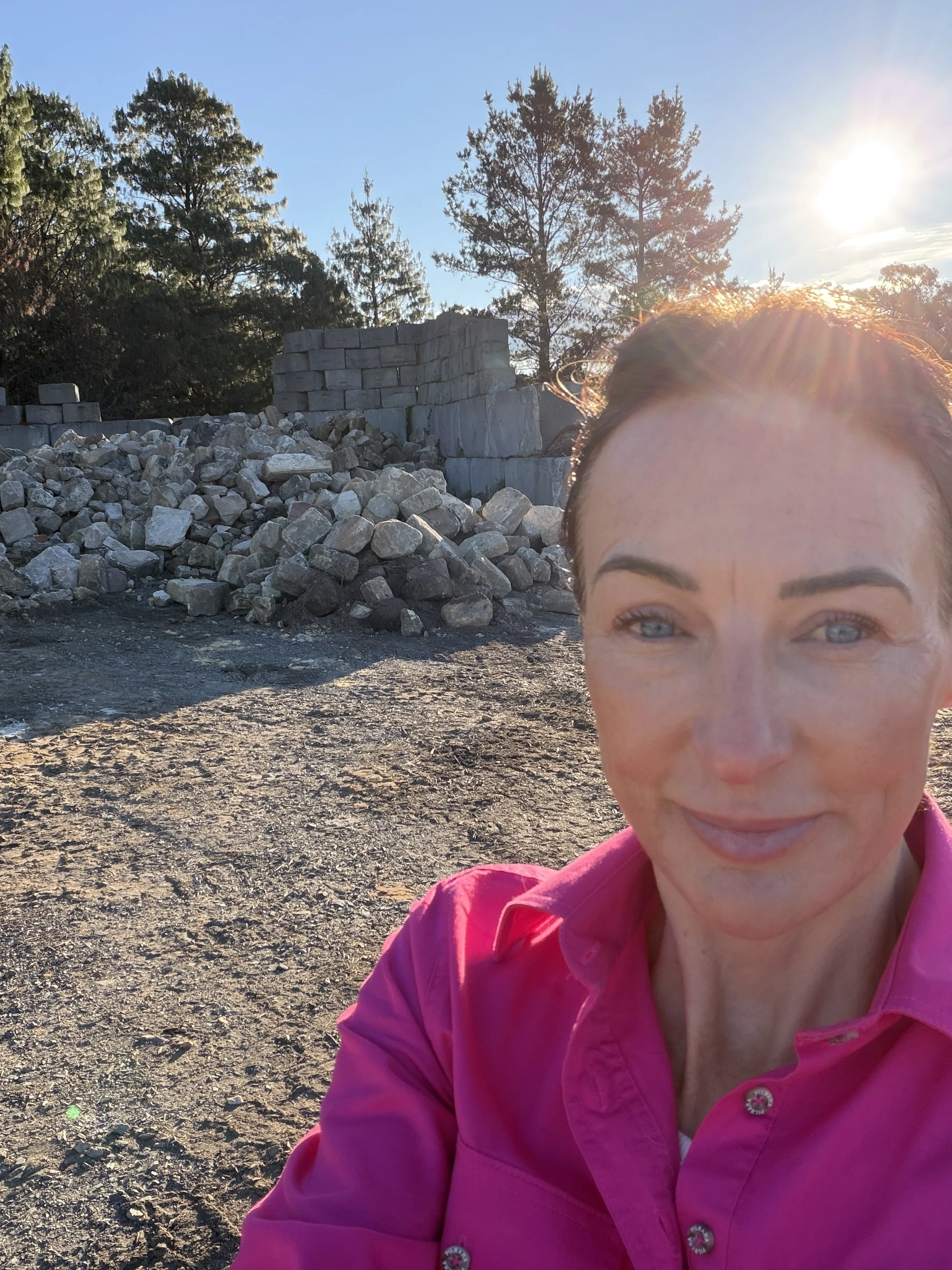 A woman in a pink jacket taking a selfie outdoors during sunset, with a backdrop of rocks, trees, and the sun shining brightly in the sky.