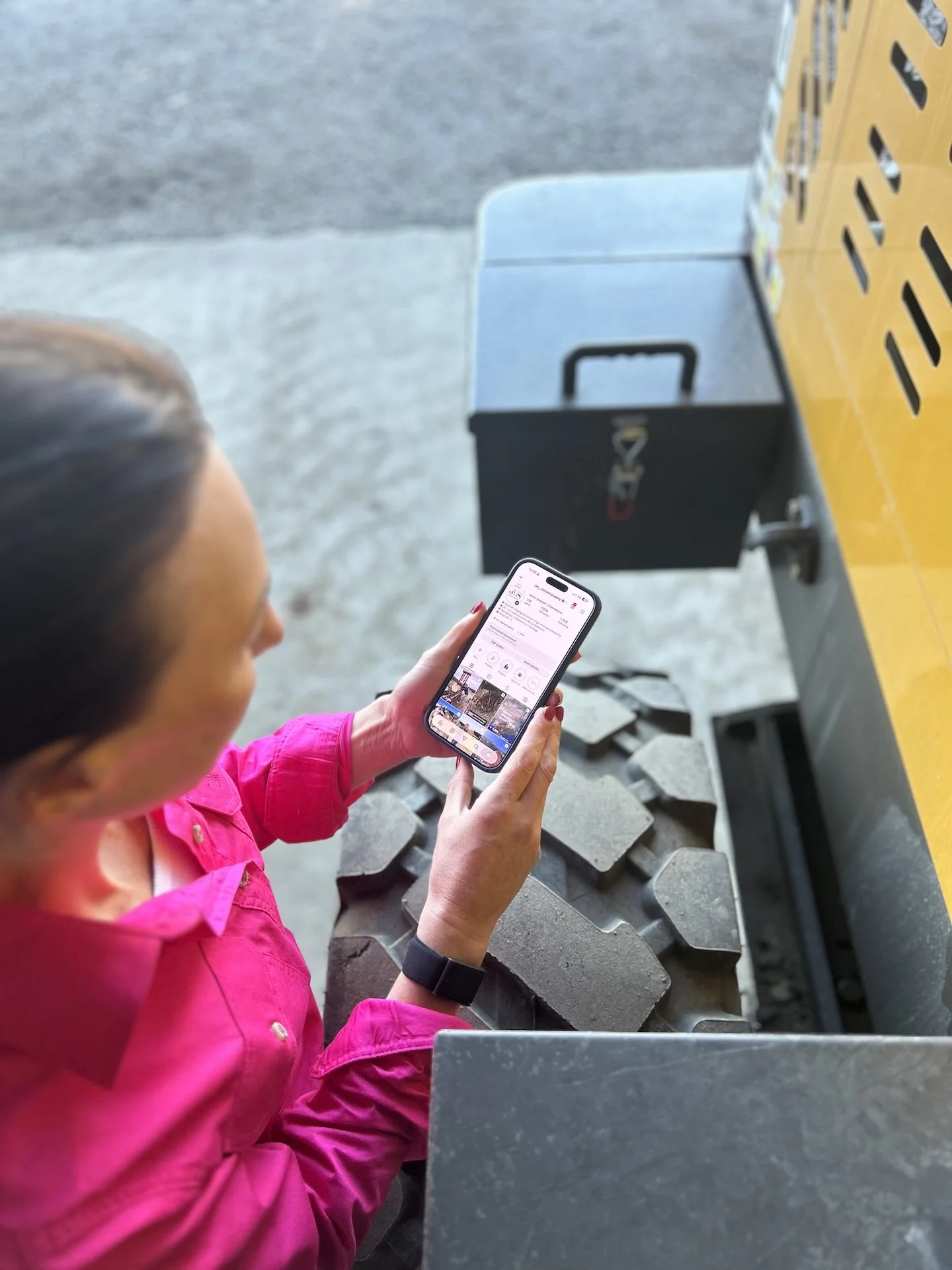 A woman in a pink shirt holding a smartphone, standing next to a yellow industrial machine with large tires.