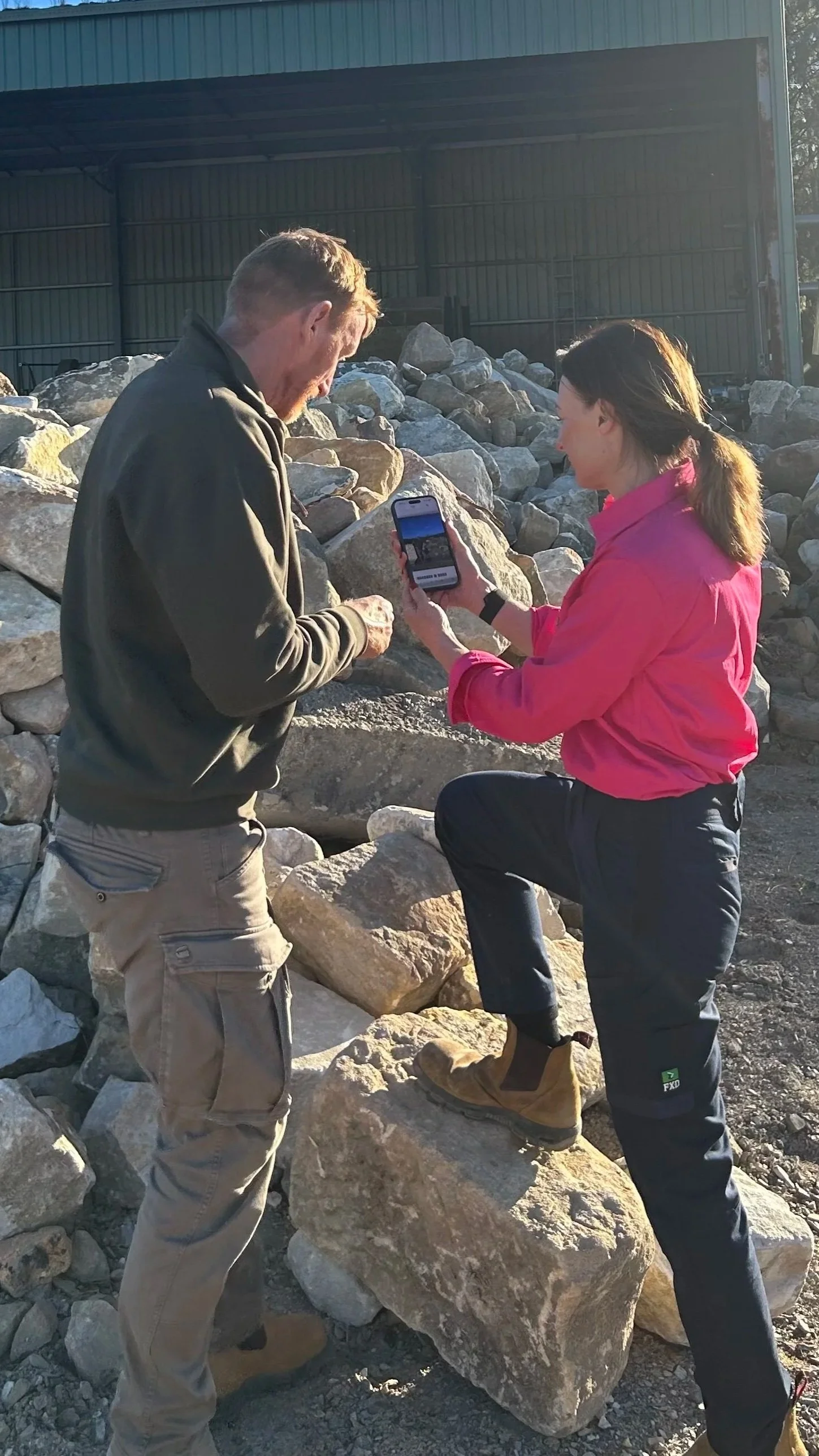 Two people, a man and a woman, at a rocky outdoor location, looking at a smartphone together. The woman is seated on a large rock, wearing a pink jacket and black pants, and the man is standing beside her, wearing a dark jacket and khaki pants.