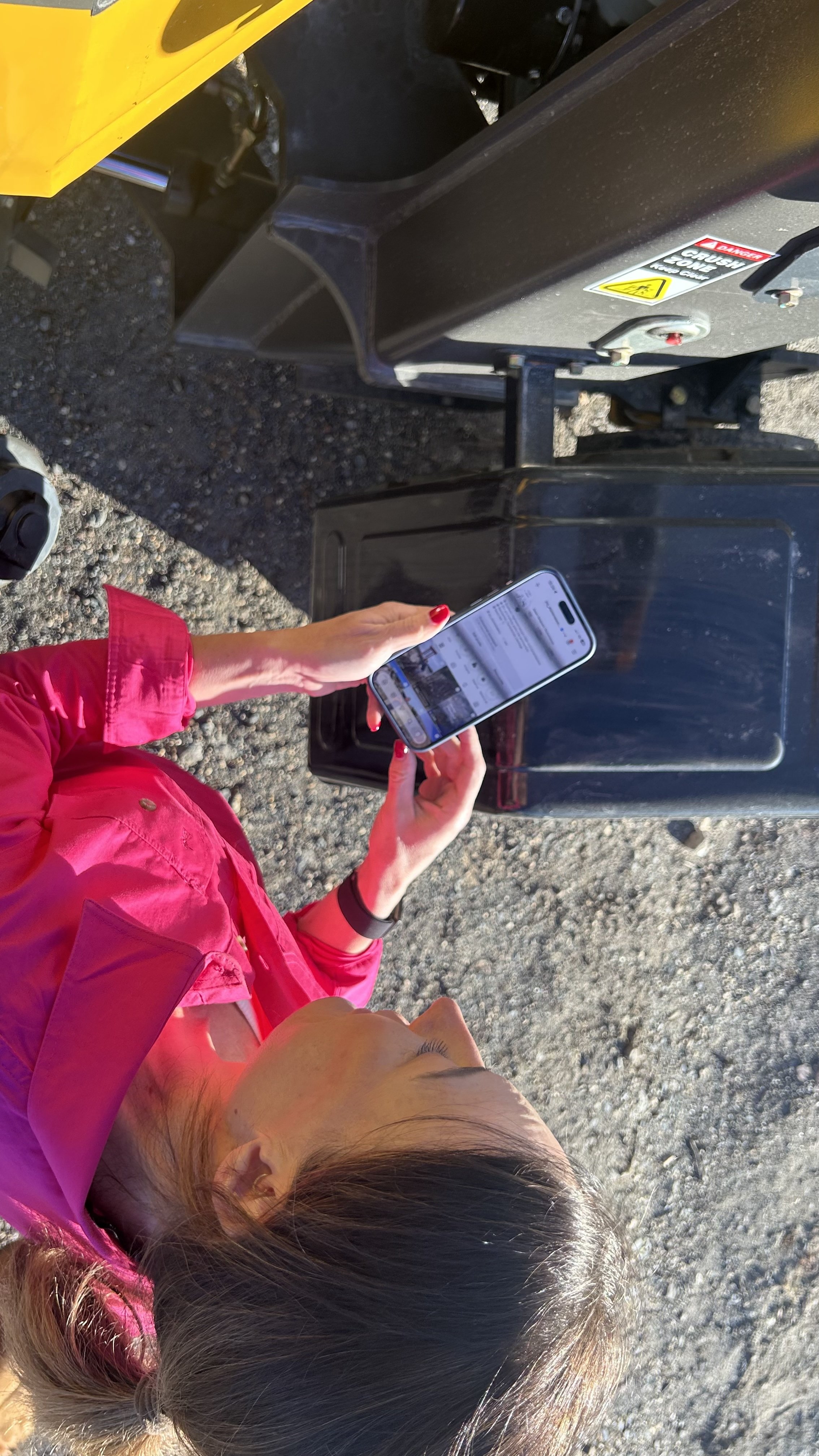 A woman with dark hair and light skin, wearing a bright pink jacket, is holding a smartphone and looking at it. She is outdoors on a gravel surface, near some industrial equipment or machinery.