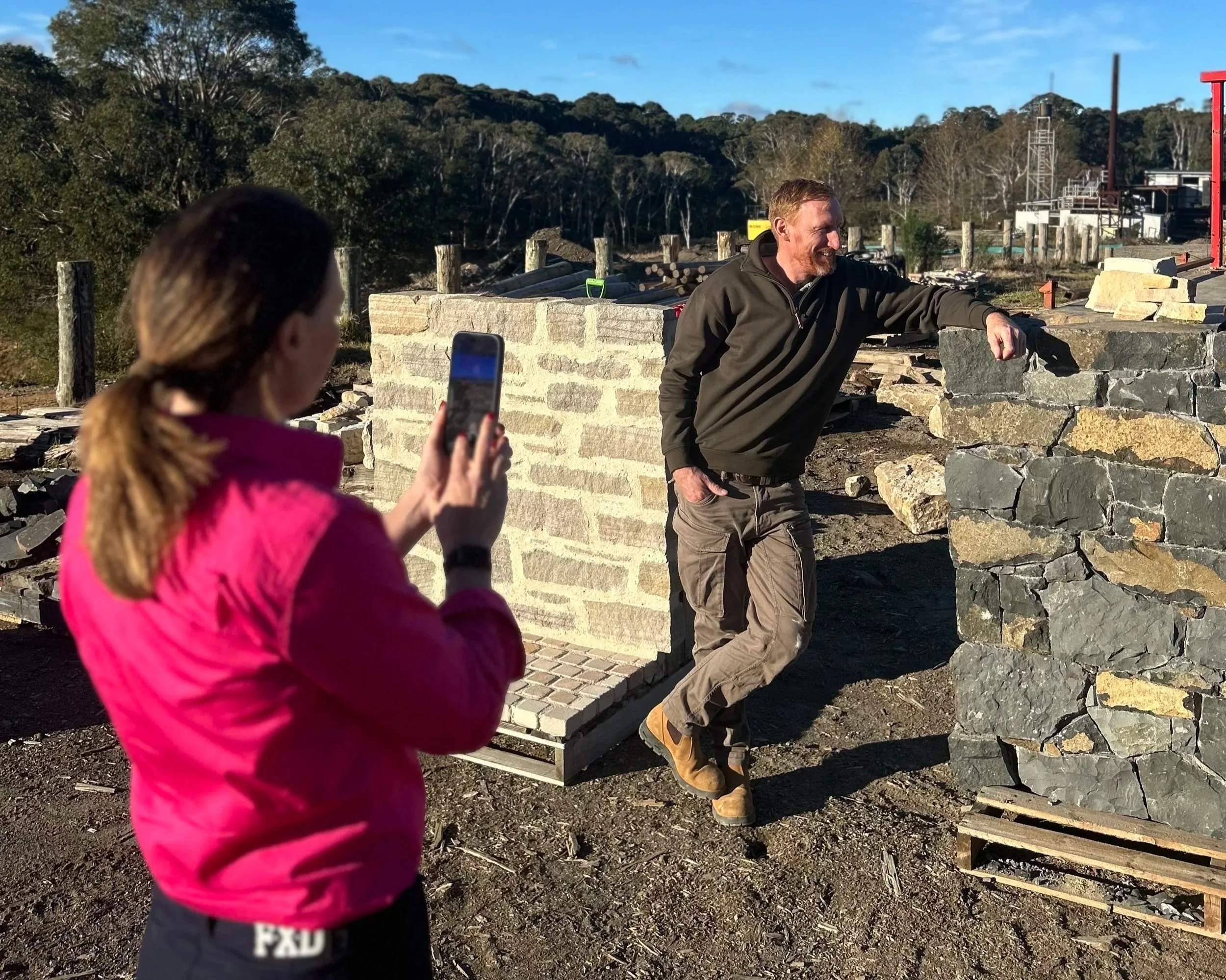 A woman in a pink jacket takes a photo of a man in outdoor work clothes, who is smiling and leaning on a stone wall while working at a construction site with trees and blue sky in the background.