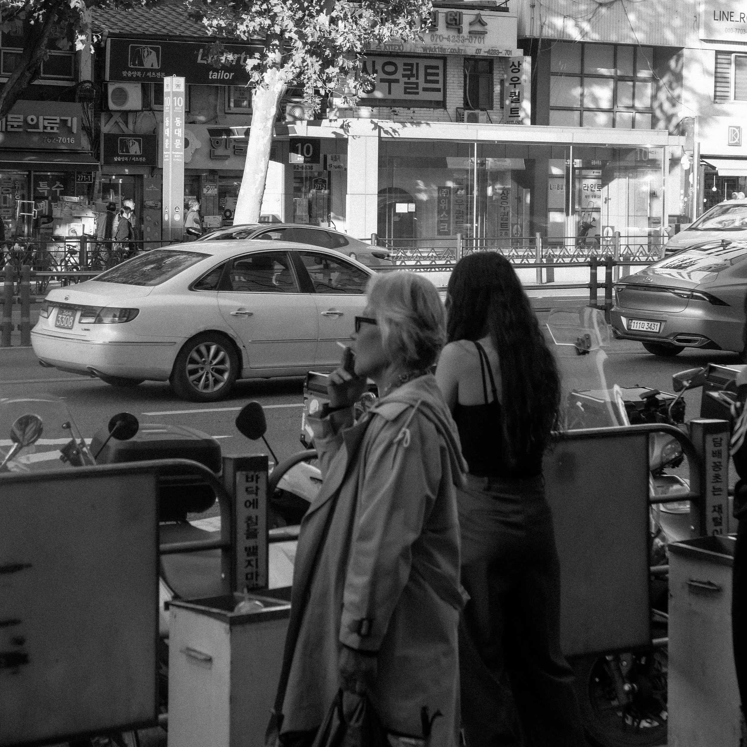 A black and white street scene with parked cars, pedestrians, and multiple storefronts with signage in Korean.