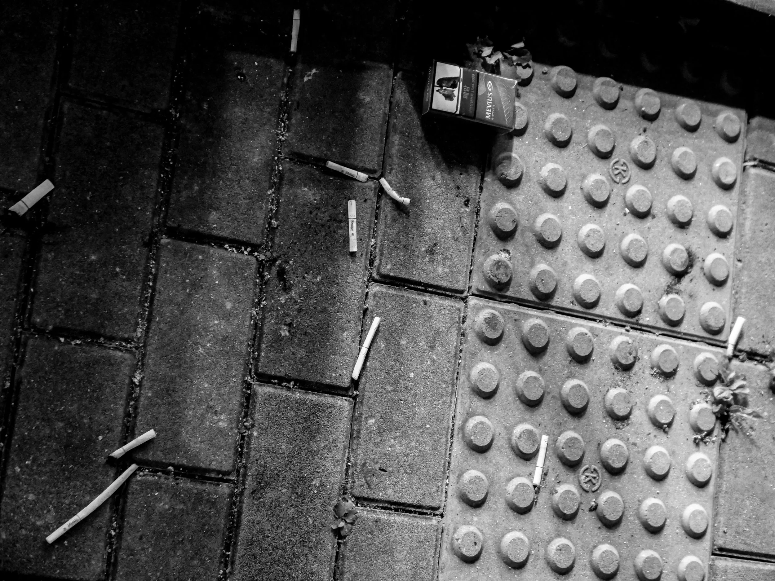 Street sidewalk with cigarette butts scattered, a faded cigarette pack, and a section of tactile paving for the visually impaired.