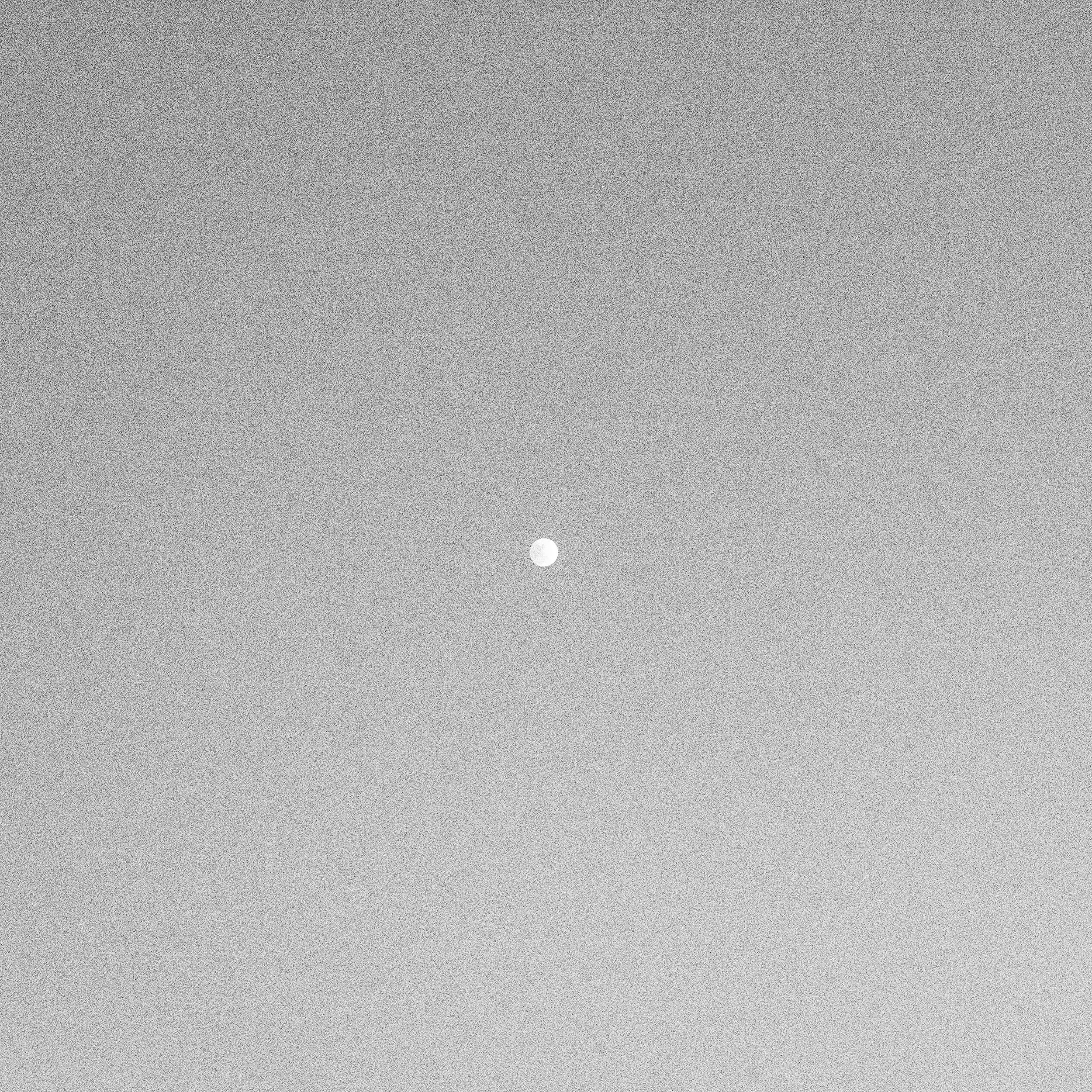 A small white object, possibly a moon or planet, is seen against a gray textured sky with a grainy appearance.