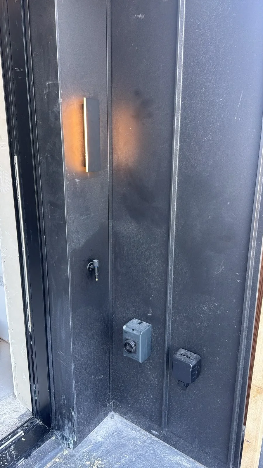 Close-up of a black metal wall with outdoor lighting, electrical box, and a water spigot.