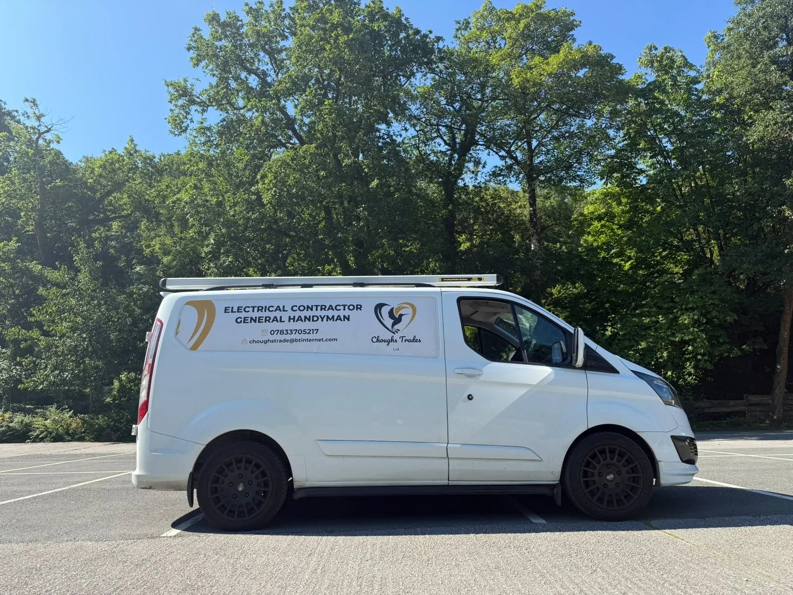 White van parked in a lot with a background of green trees and a clear blue sky. The van has signage indicating electrical contractor and handyman services.