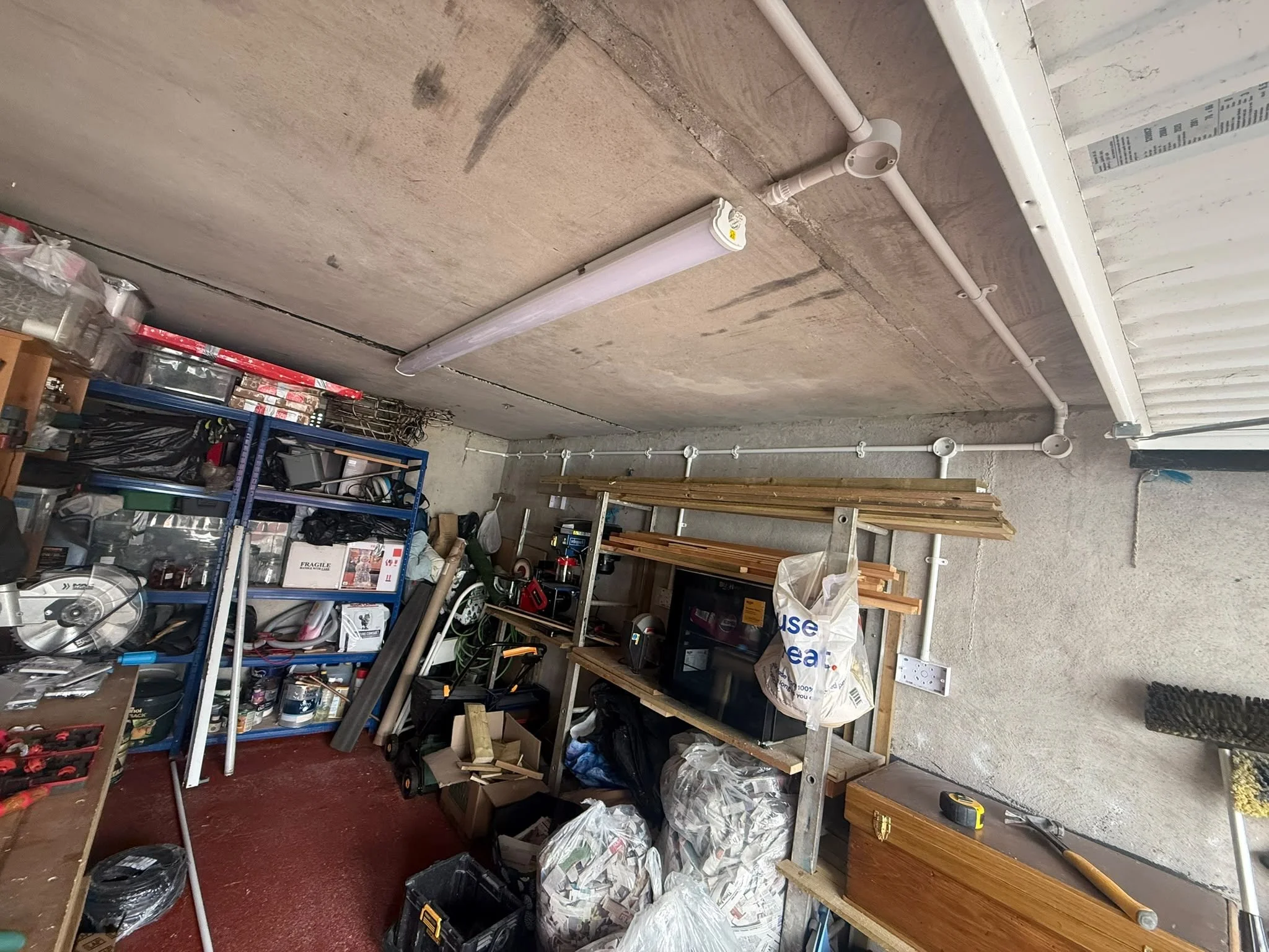 View of a cluttered garage workspace with shelves filled with tools, boxes, and supplies, and an unfinished wall with visible pipes and utility connections.
