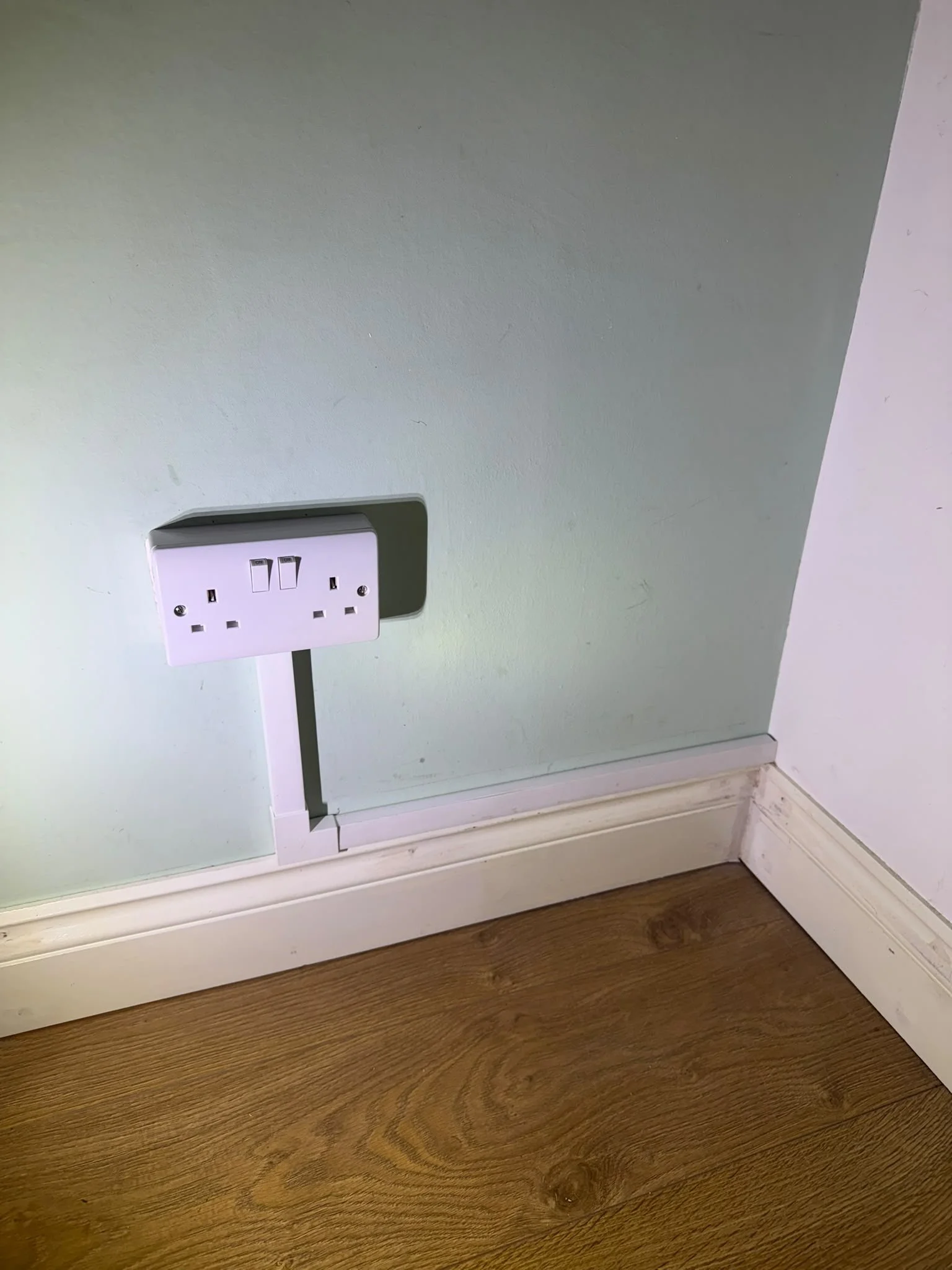 A wall outlet with a white cover plate, connected to an extension cord, near a wooden floor and a white baseboard.