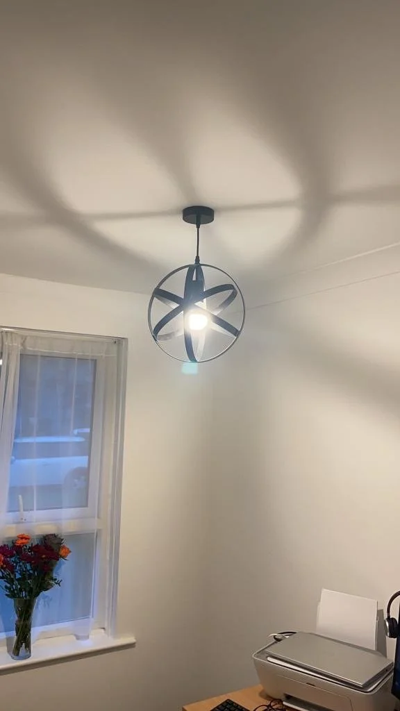 A modern black star-shaped pendant light fixture hanging from a white ceiling. The light casts star-shaped shadows on the ceiling. There is a white window with sheer curtains and a vase of colorful flowers on the windowsill. A corner of a desk with a closed laptop and a pair of headphones is visible.