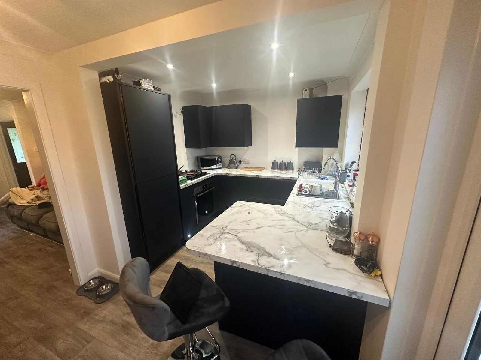 Modern kitchen with black cabinets, white marble countertops, and various appliances including a microwave, stove, and dishwasher. There is a marble breakfast bar and a gray upholstered bar stool.