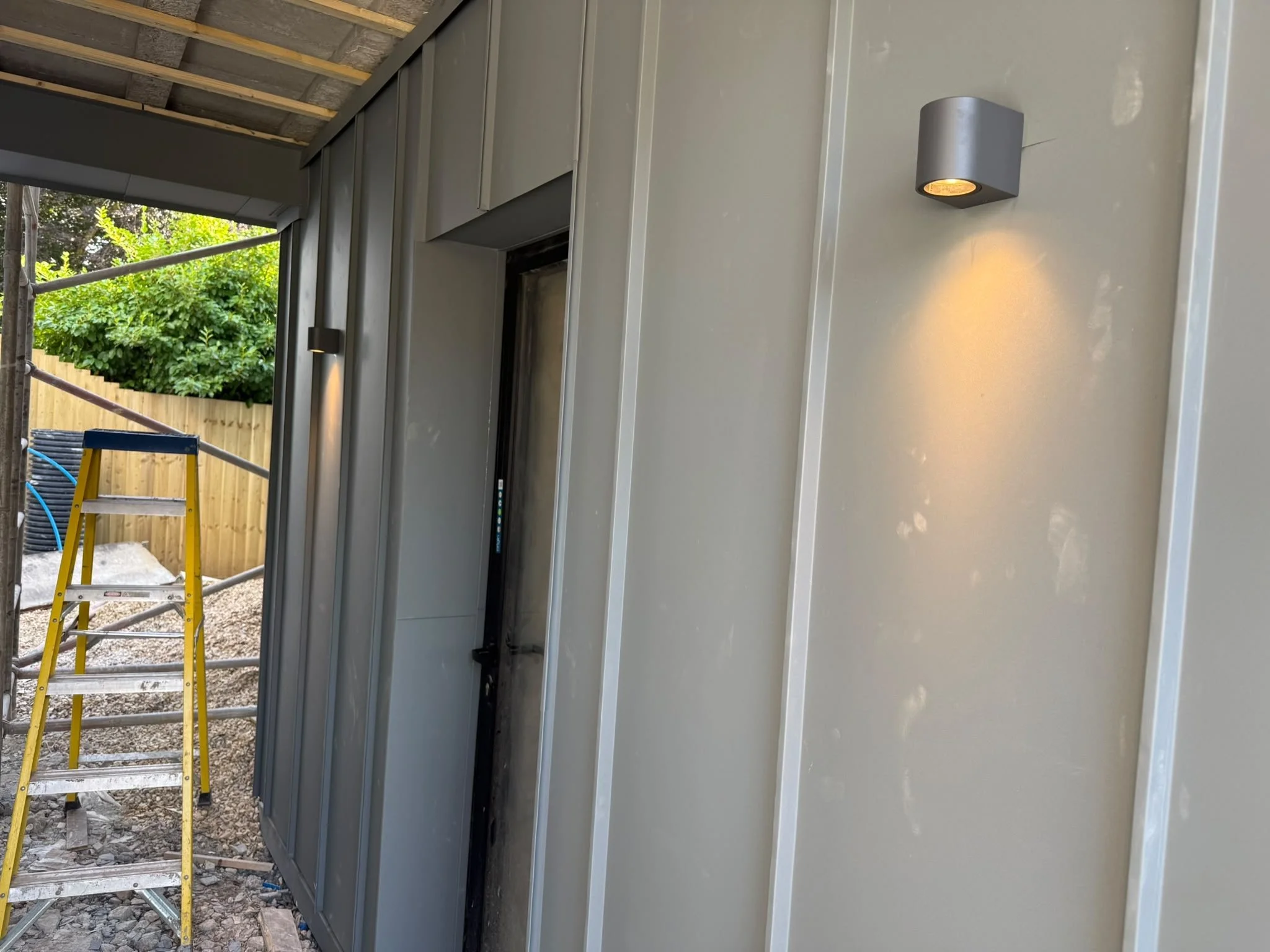 Construction site showing a partially built structure with gray metal siding on the exterior wall and a small outdoor wall light. There is a yellow step ladder and scaffolding in the background with trees and a wooden fence.