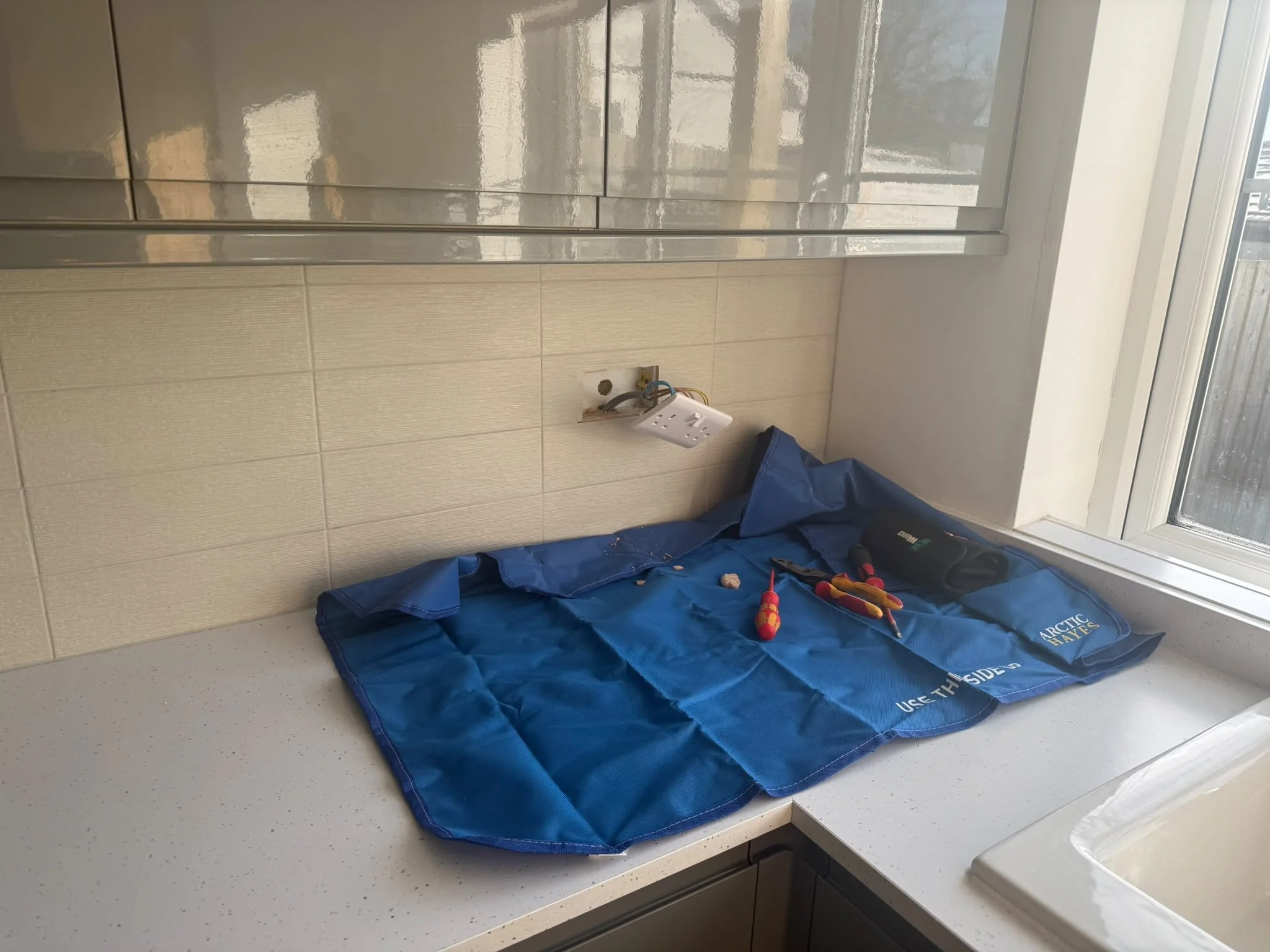 A kitchen countertop with a blue moving blanket, various tools, and an opened electrical outlet in the wall. The background shows a window and a tile wall.