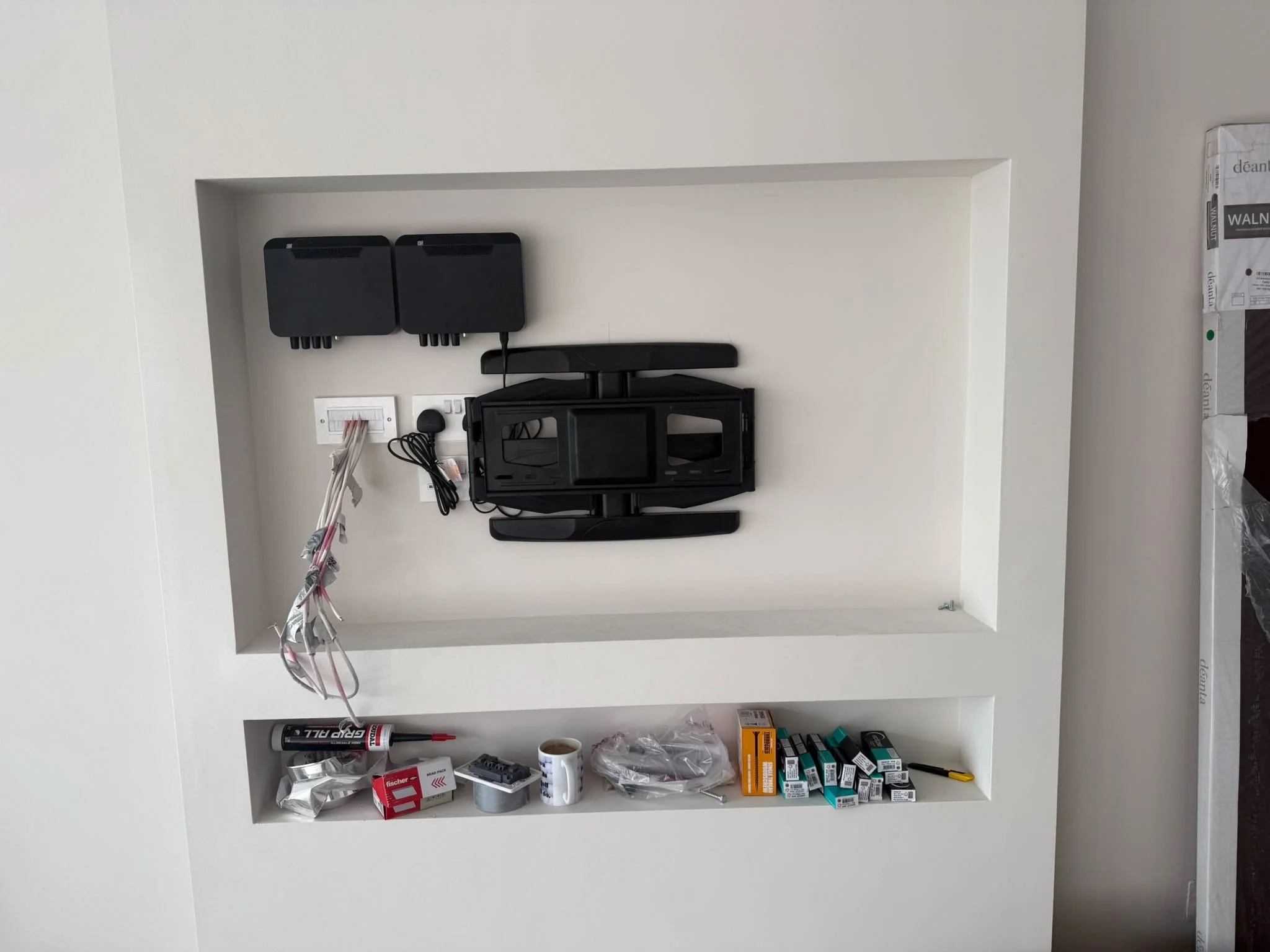 A small built-in white wall shelf containing various tools and supplies including adhesive tape, glue, a toolbox, small boxes, and miscellaneous hardware objects, with an exposed wiring bundle hanging from the wall.