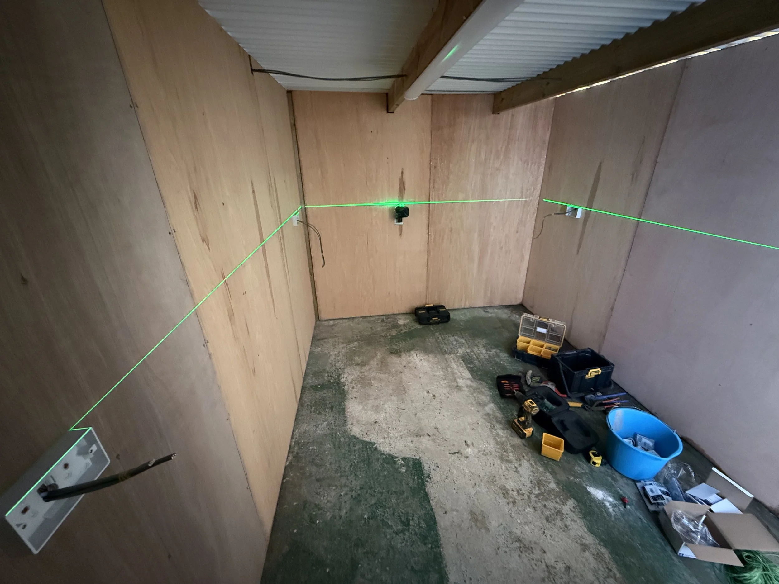 Room with unfinished wooden walls, tools, a blue bucket, black toolbox, and a green laser level projecting lines on the walls and ceiling.