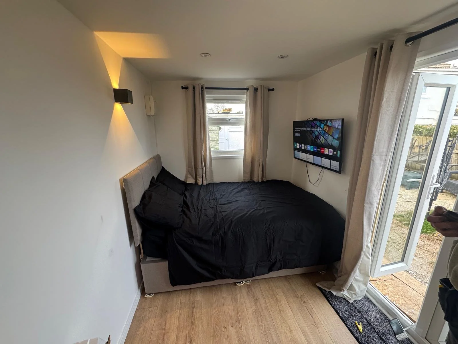 Small bedroom with a bed covered in black bedding, a wall-mounted TV, a window with beige curtains, and a glass door leading outside, with a glimpse of a backyard and patio.