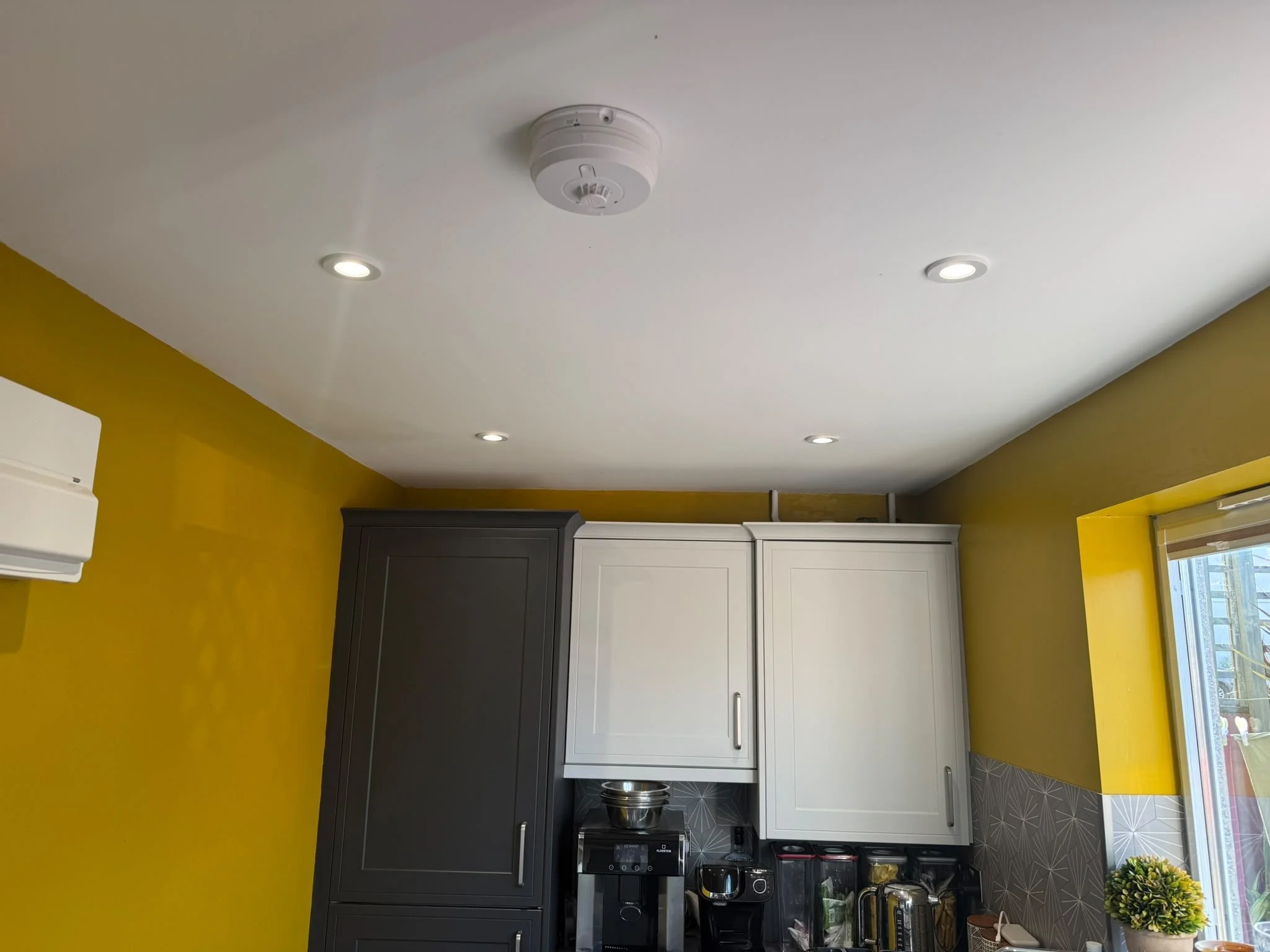 Kitchen with white and black cabinets, yellow walls, overhead ceiling lights, and a window with blinds.