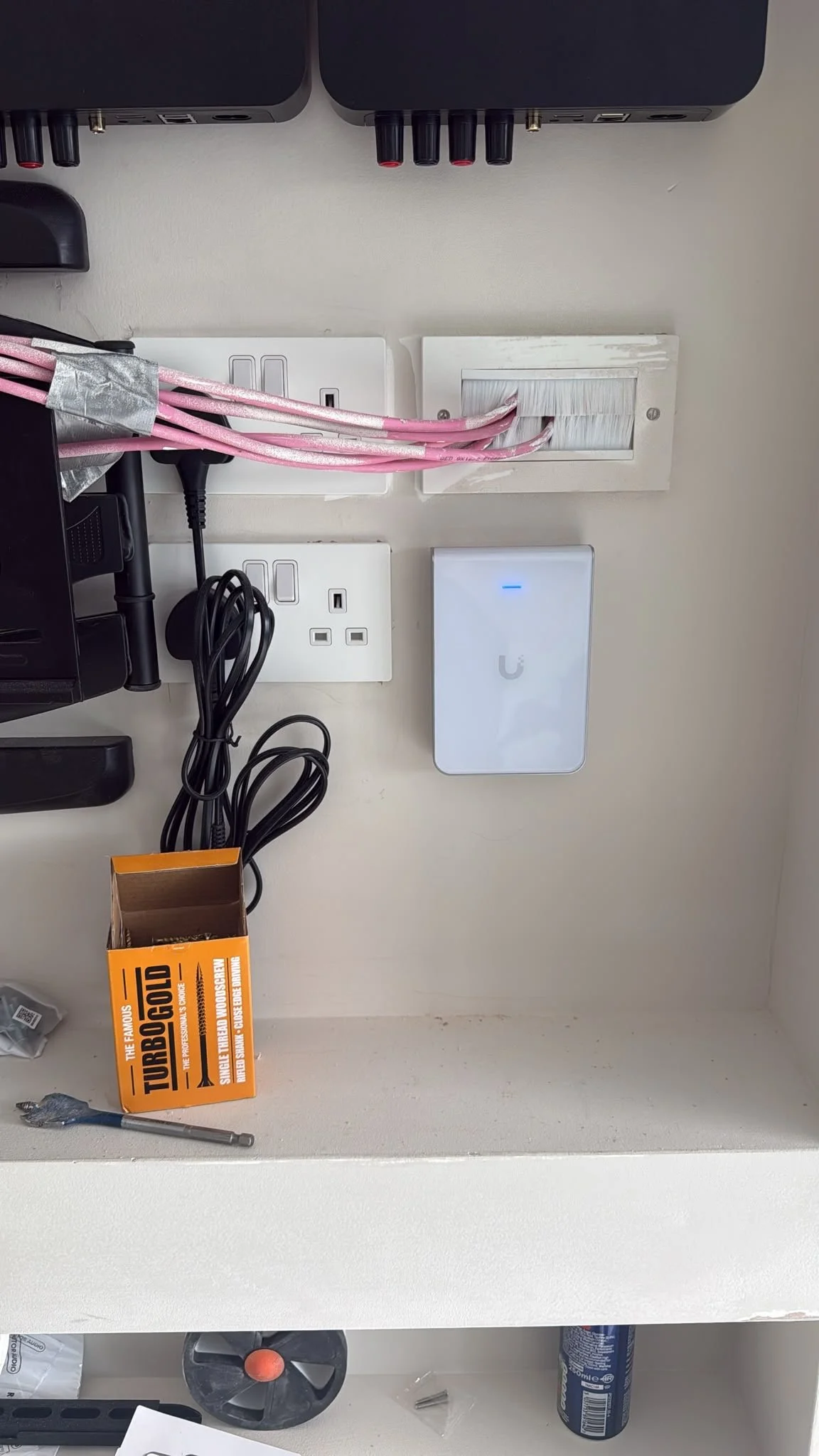 Desk with various electronic devices and tools, including a white Wi-Fi extender, power outlets, a power strip with plugged-in cords, a small open box of screws, a screwdriver, and some packaging materials.