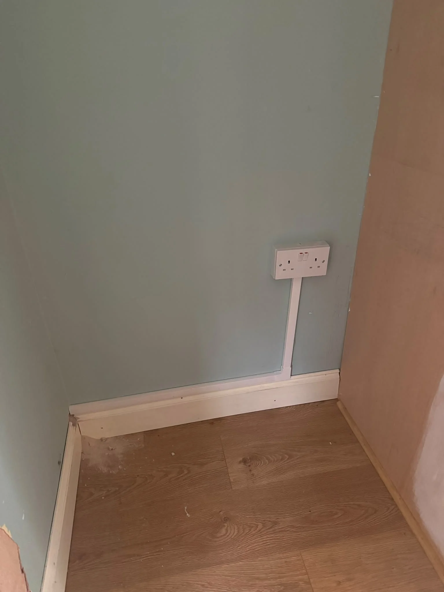 Corner of a room with a light green wall, a wooden floor, and a white baseboard. An electrical outlet is mounted on the wall near the floor, with a white cable connected to it.