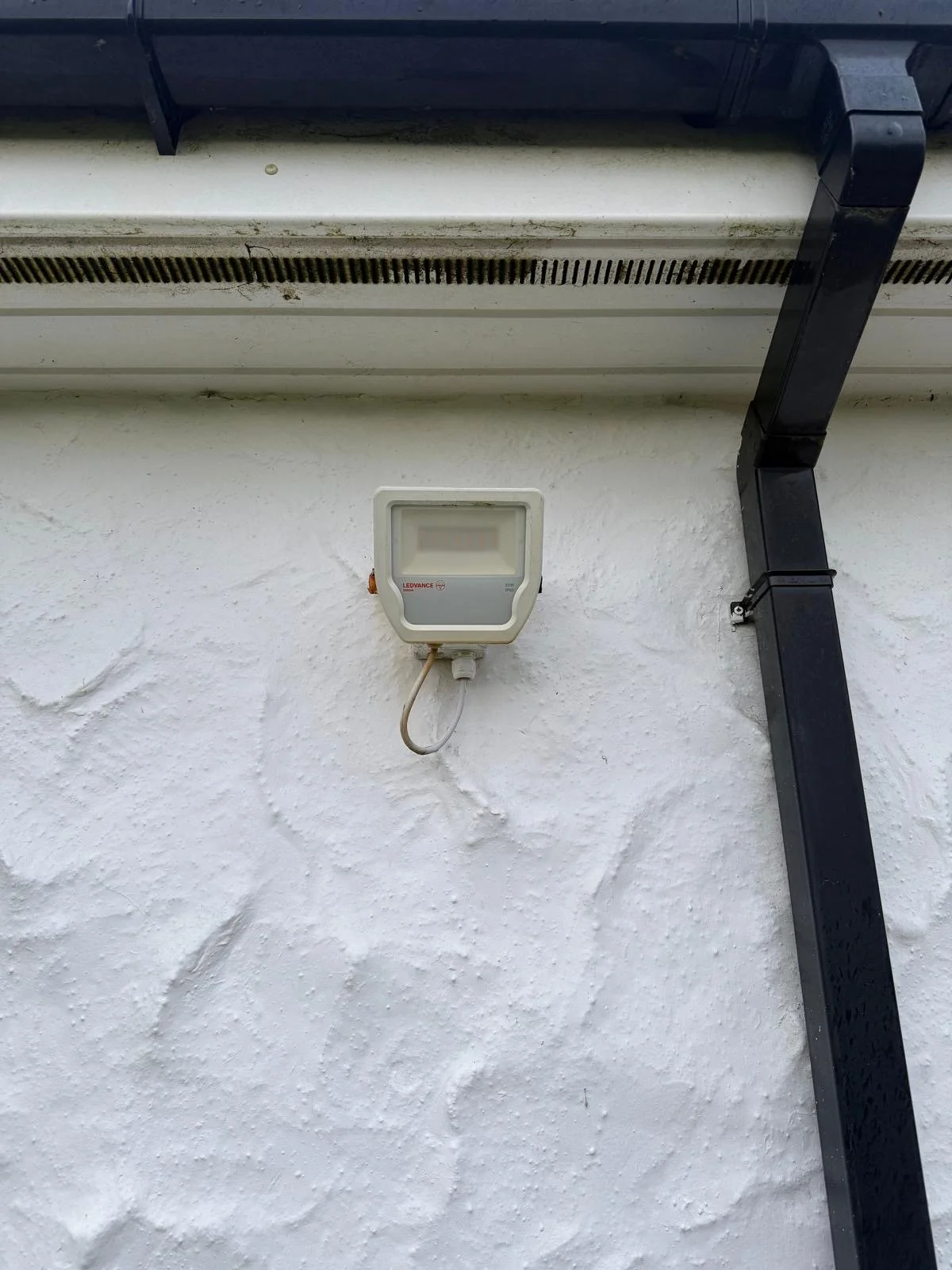 Outdoor white textured wall with a small electronic device, possibly a garage door opener or sensor, attached to it. Above the device, there is a black pipe or conduit running vertically along the wall and a vent with horizontal slats near the ceiling.