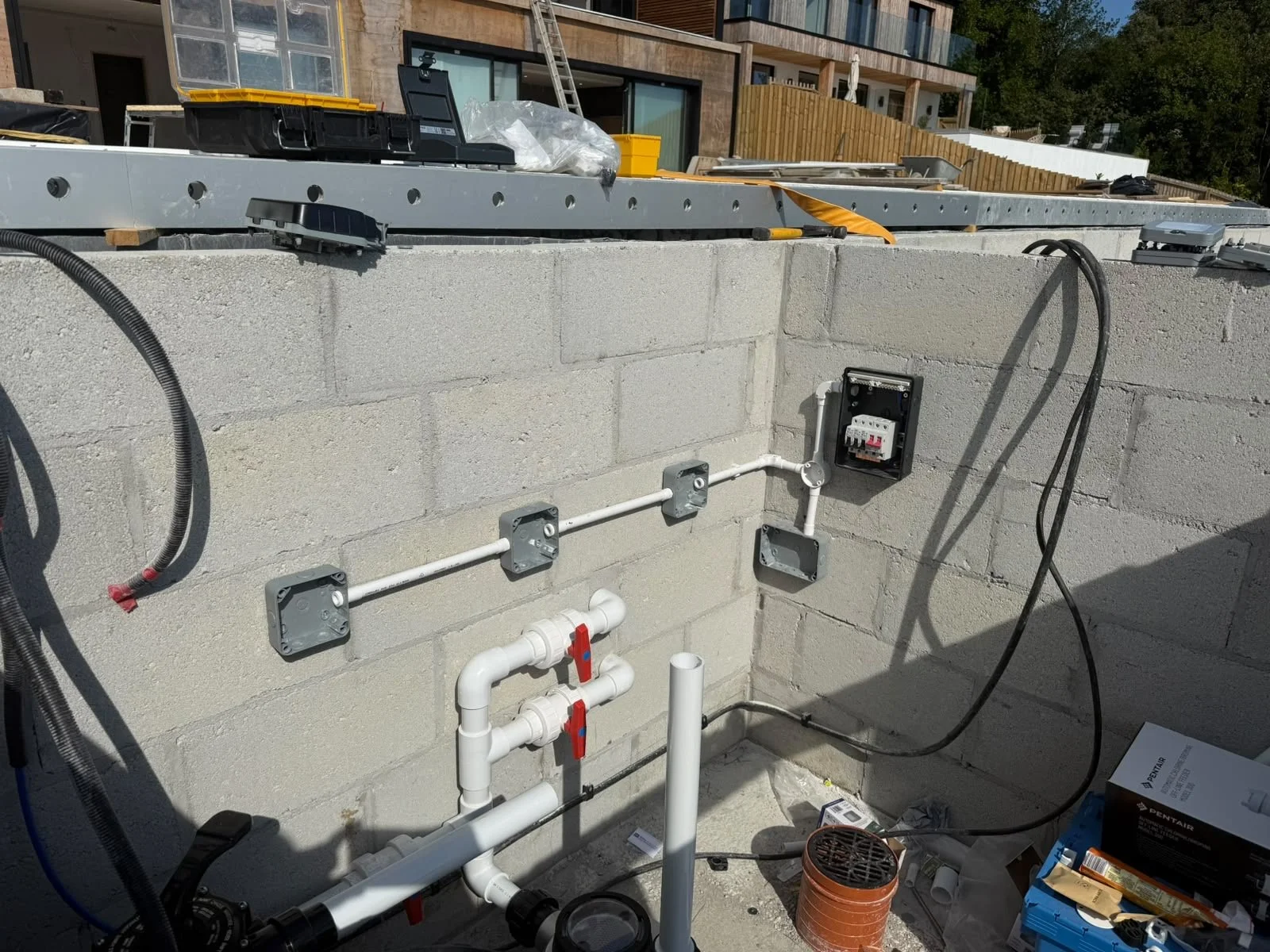 Construction site with electrical wiring and plumbing installed on a concrete block wall, with tools and materials on the ground.