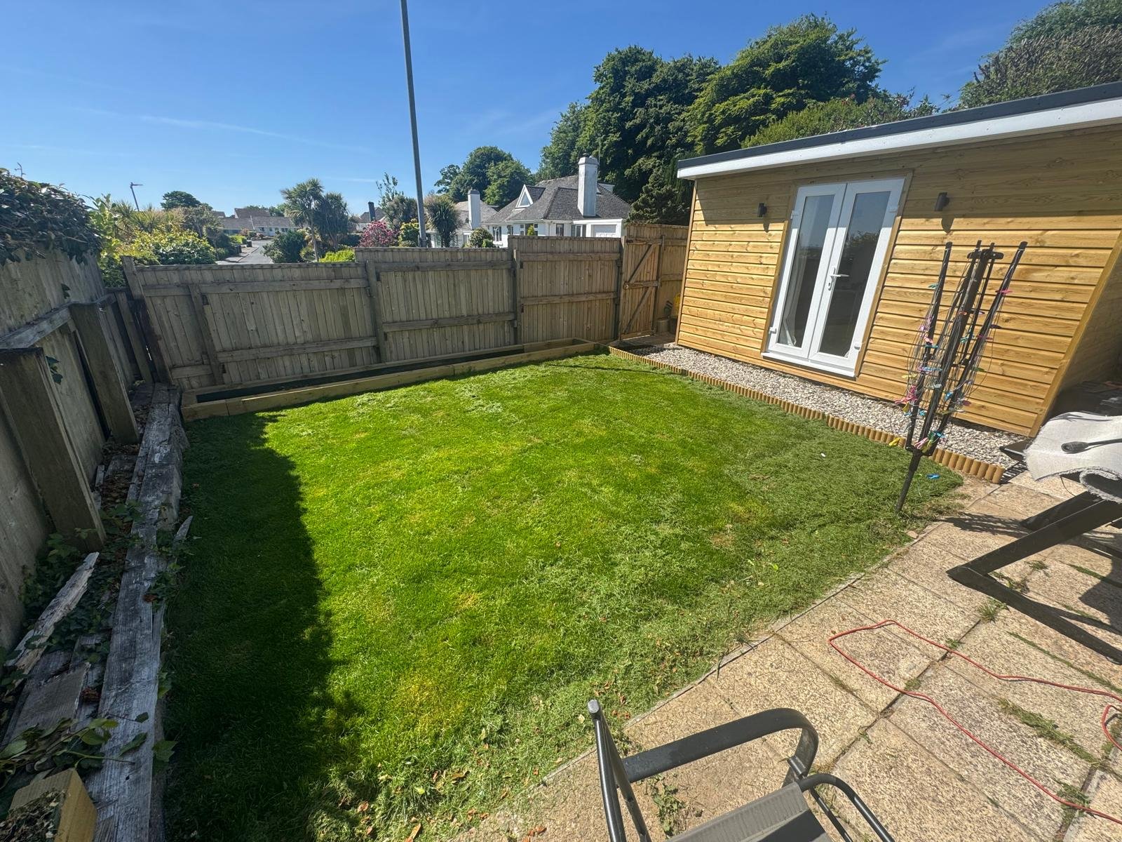 Backyard with green lawn, wooden fence, small wooden shed with glass doors, and outdoor furniture under a sunny sky.