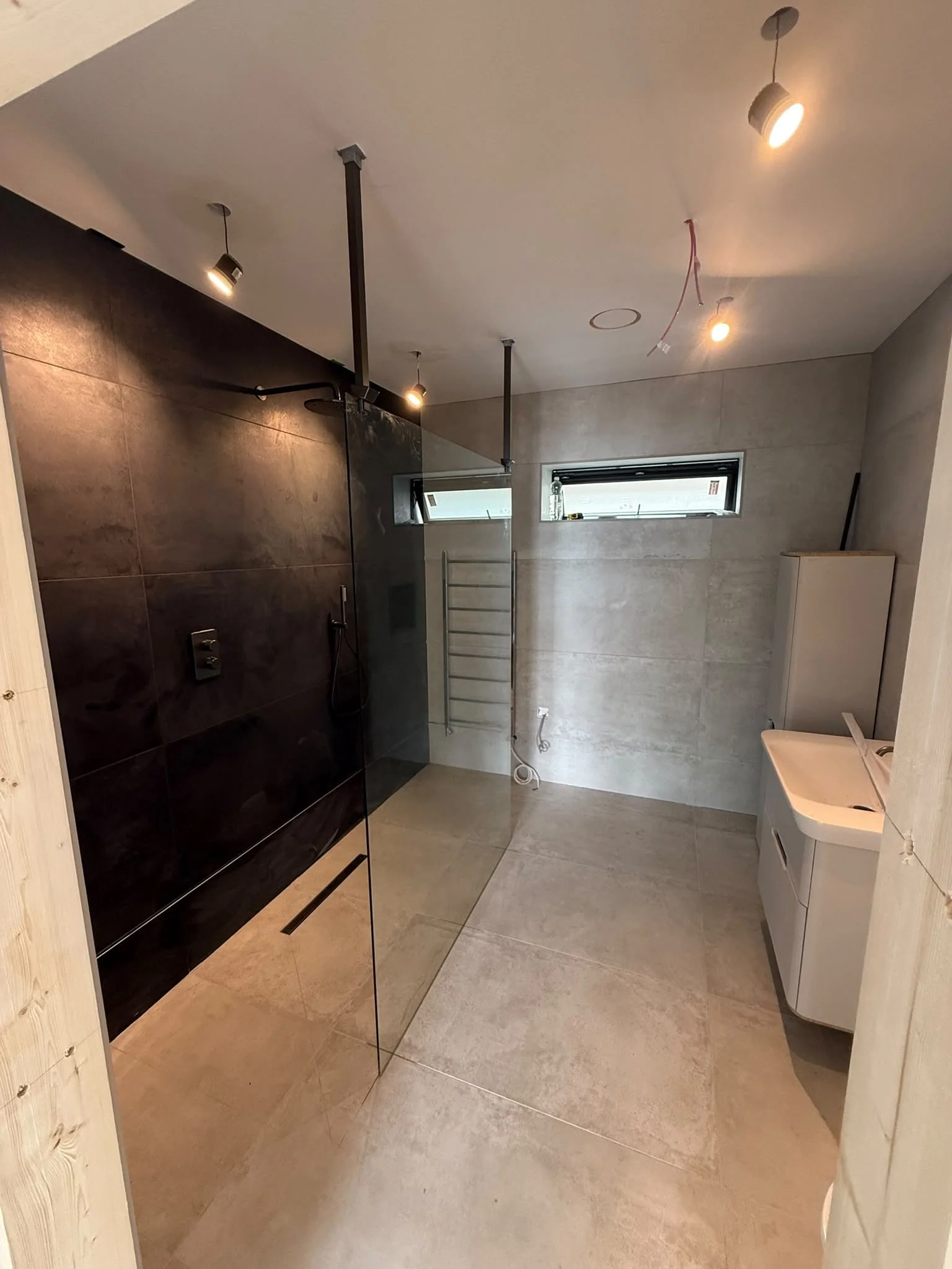A modern bathroom with beige tiled flooring and walls. There is a wall-mounted shower with a black tiled wall and a glass partition. The ceiling has multiple recessed and track lights. A small window is near the ceiling. On the right, there is a white sink cabinet with a digital sensor faucet. A wooden panel partially surrounds the area.