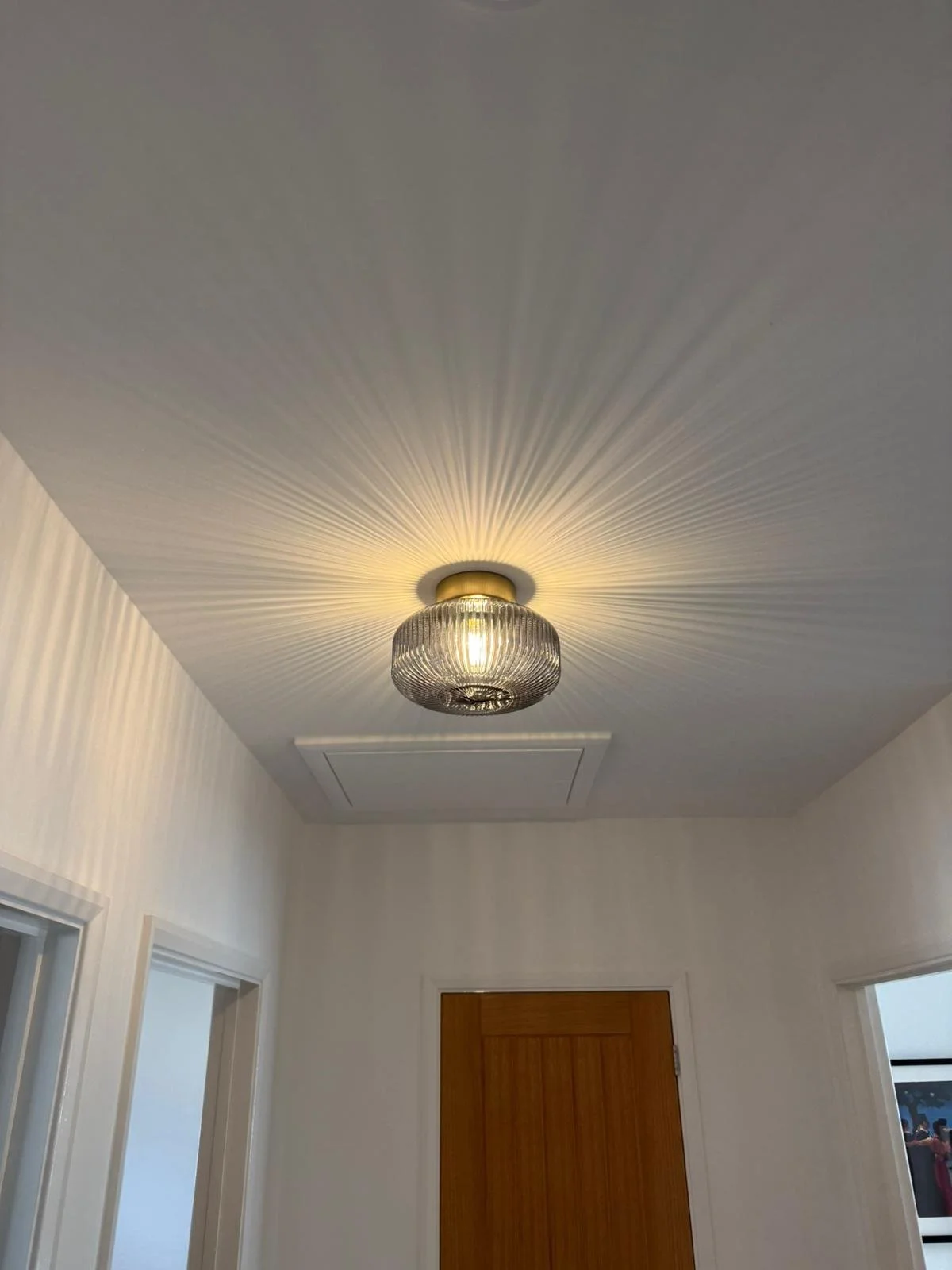 A ceiling light fixture with a glass or crystal cover casting radial light patterns on the white ceiling in a hallway with white walls and a wooden door.