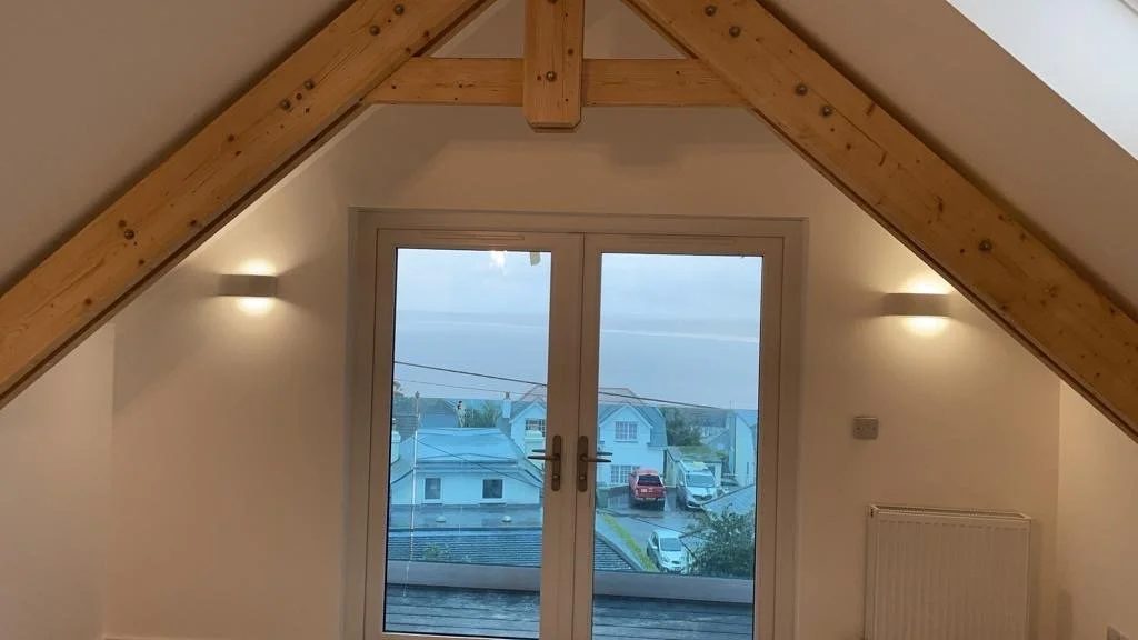 View of houses and parked cars seen through glass doors in an attic room with sloped ceiling and wooden beams.
