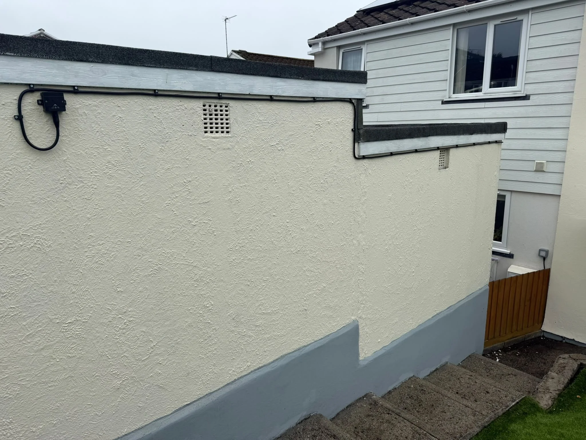 Exterior view of a house with a textured beige wall, a small vent, and a black cable conduit running along the wall. There is a window on the house, and part of a wooden fence is visible at the bottom right.
