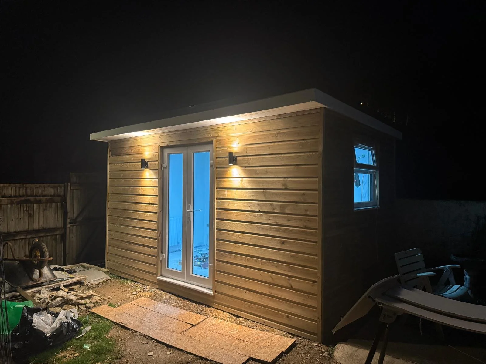 Small wooden shed with glass double doors illuminated by exterior lights, night scene, with a window showing a blue interior light, garden furniture and building materials nearby.