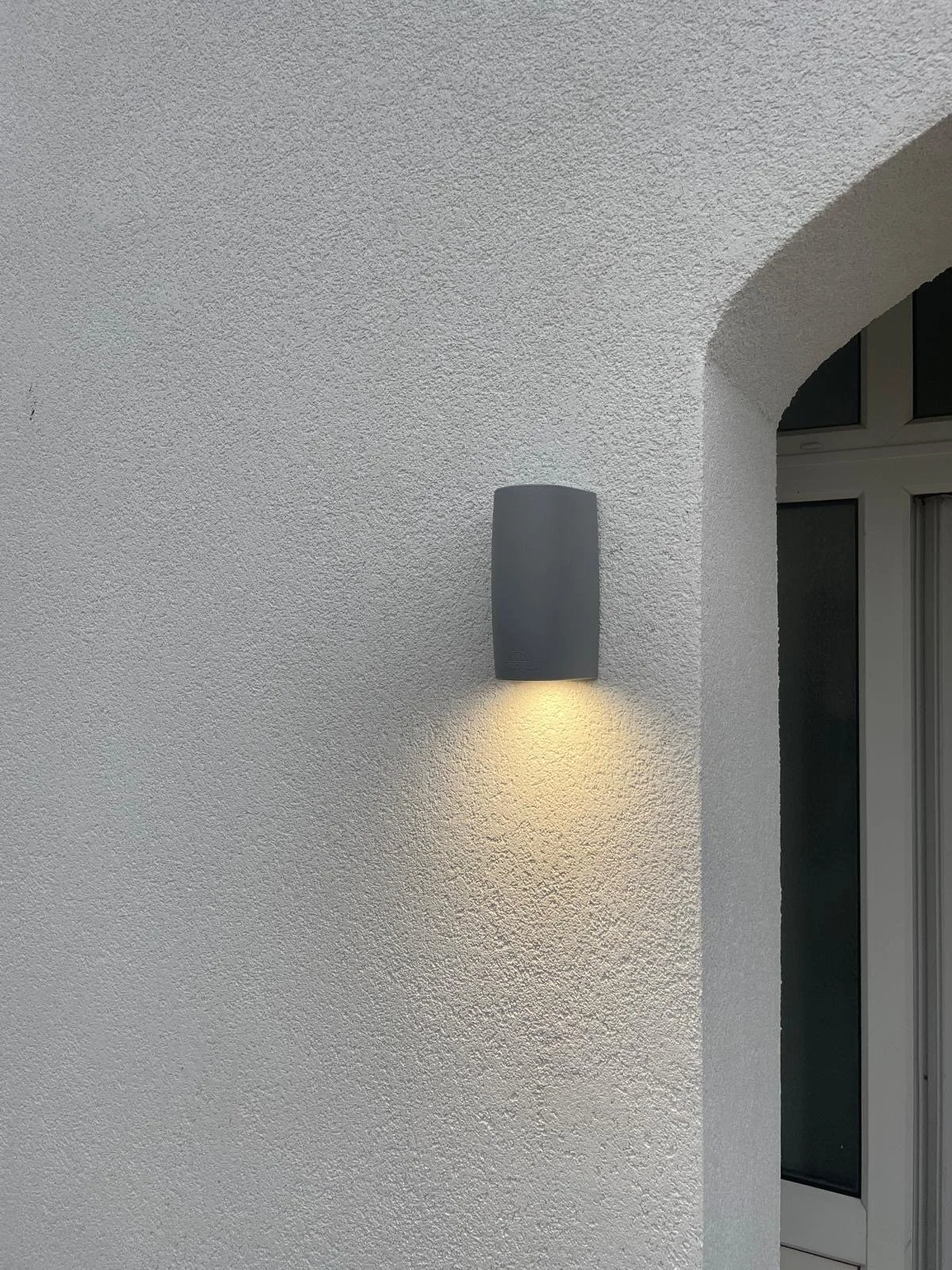 A textured white wall with a gray outdoor wall-mounted light fixture casting a warm yellow light downward. Part of a doorway or window with a white frame visible on the right side.