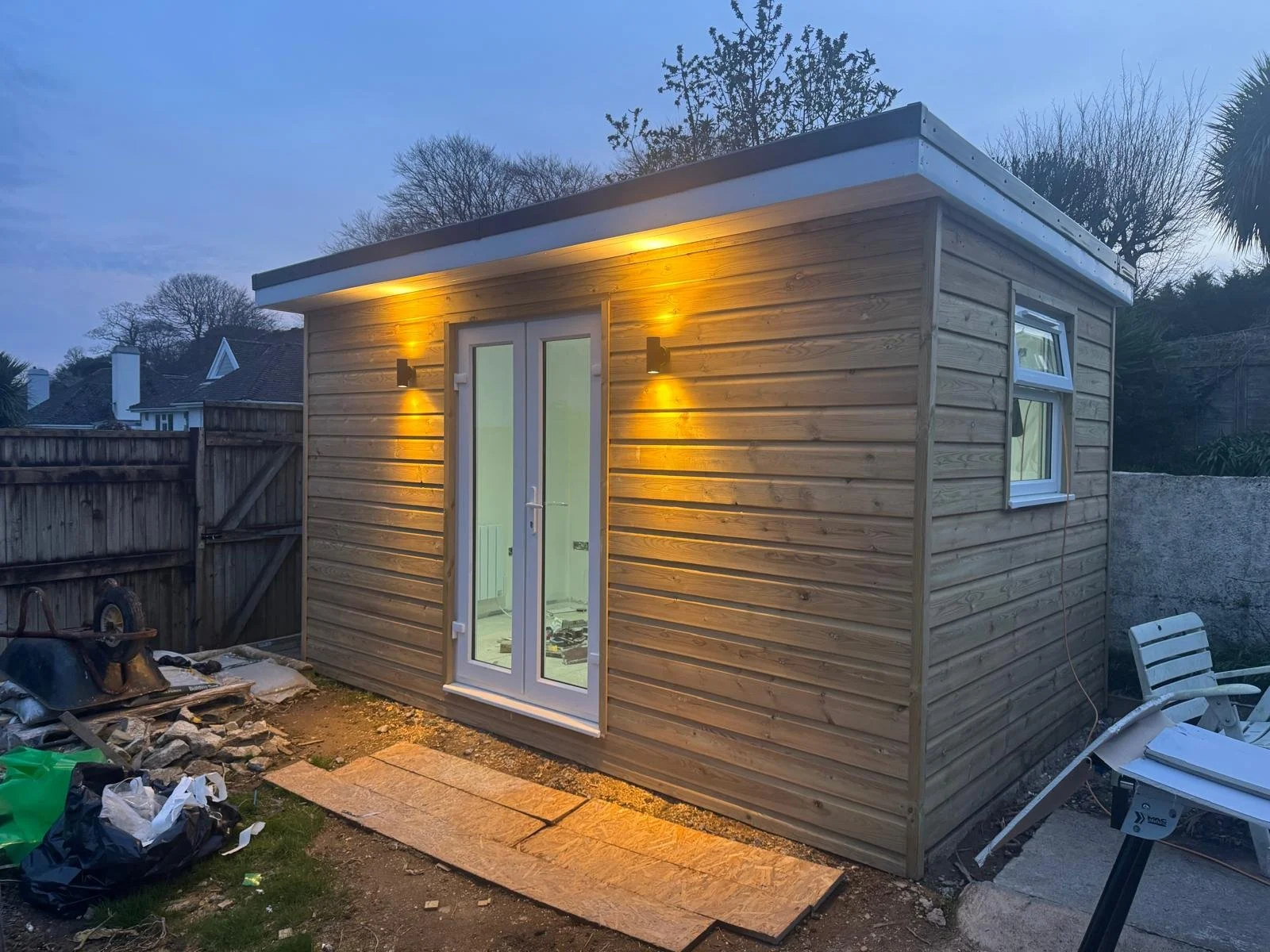 A small wooden shed or cabin under construction with exterior lighting, surrounded by construction materials and tools, and nearby outdoor furniture.