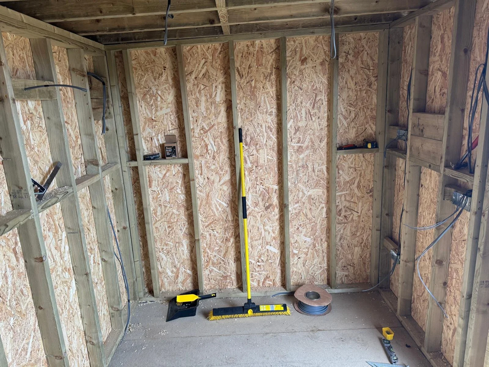 Interior of a room under construction with exposed wooden framing, electrical wiring, and tools such as broom, measuring tape, and level on the floor.