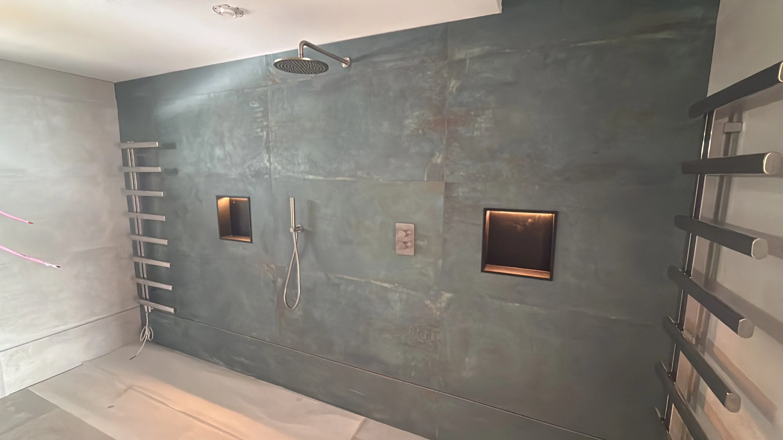 A modern shower area with a sleek, gray stone wall, ceiling-mounted showerhead, a handheld shower wand, two built-in niche shelves with lighting, and a black ladder-style towel rack on the right side.