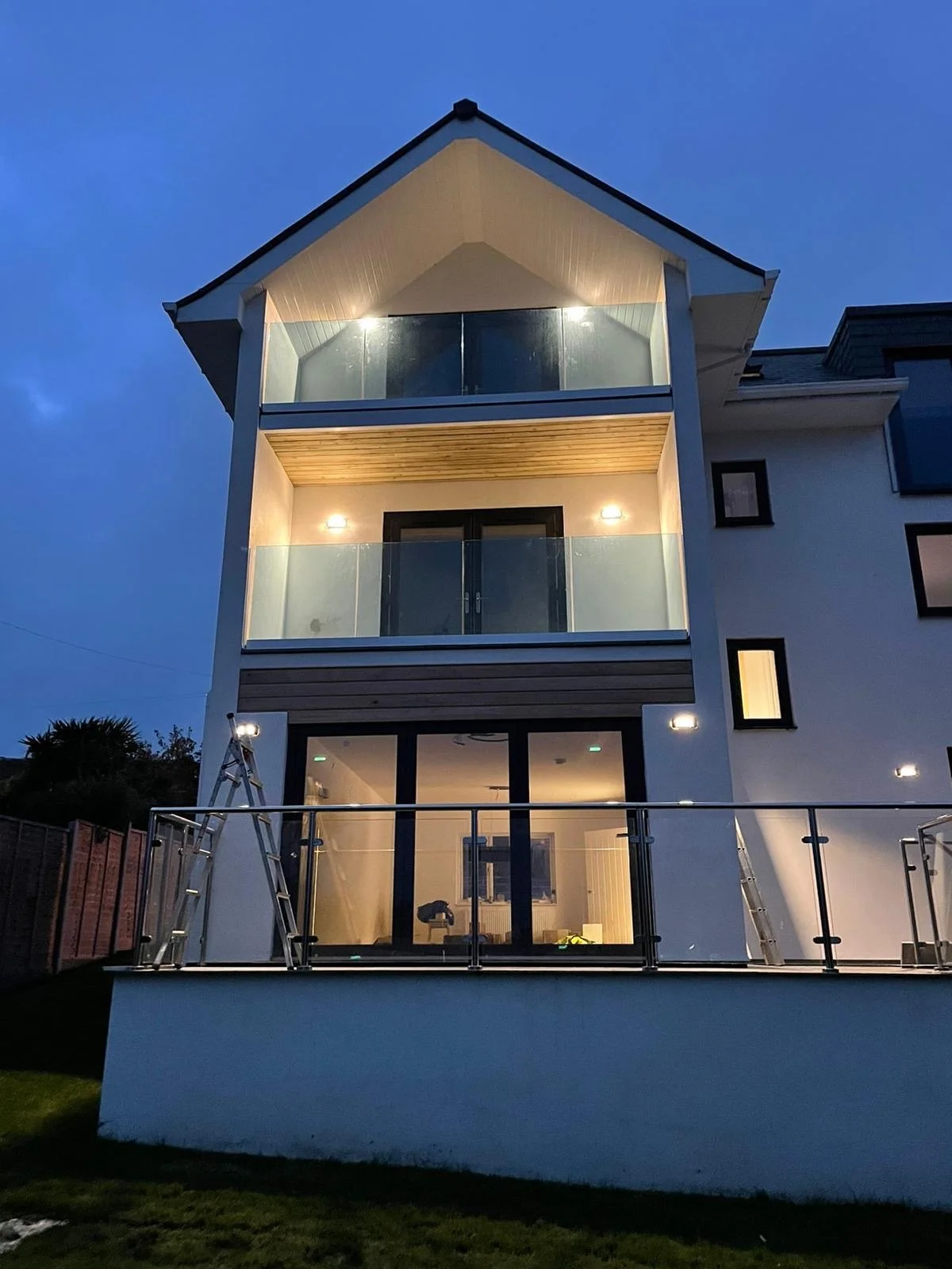 Modern three-story house with large glass windows and balconies, illuminated at dusk.