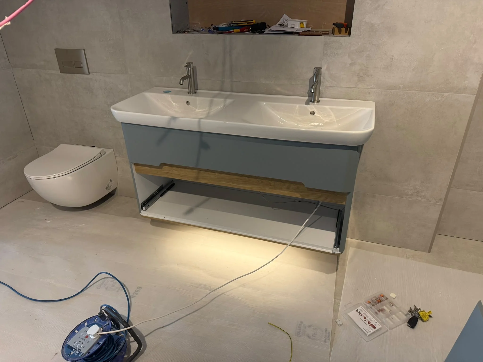 A bathroom with a dual sink vanity, a wall-mounted toilet, and construction tools scattered around, indicating ongoing installation or renovation.