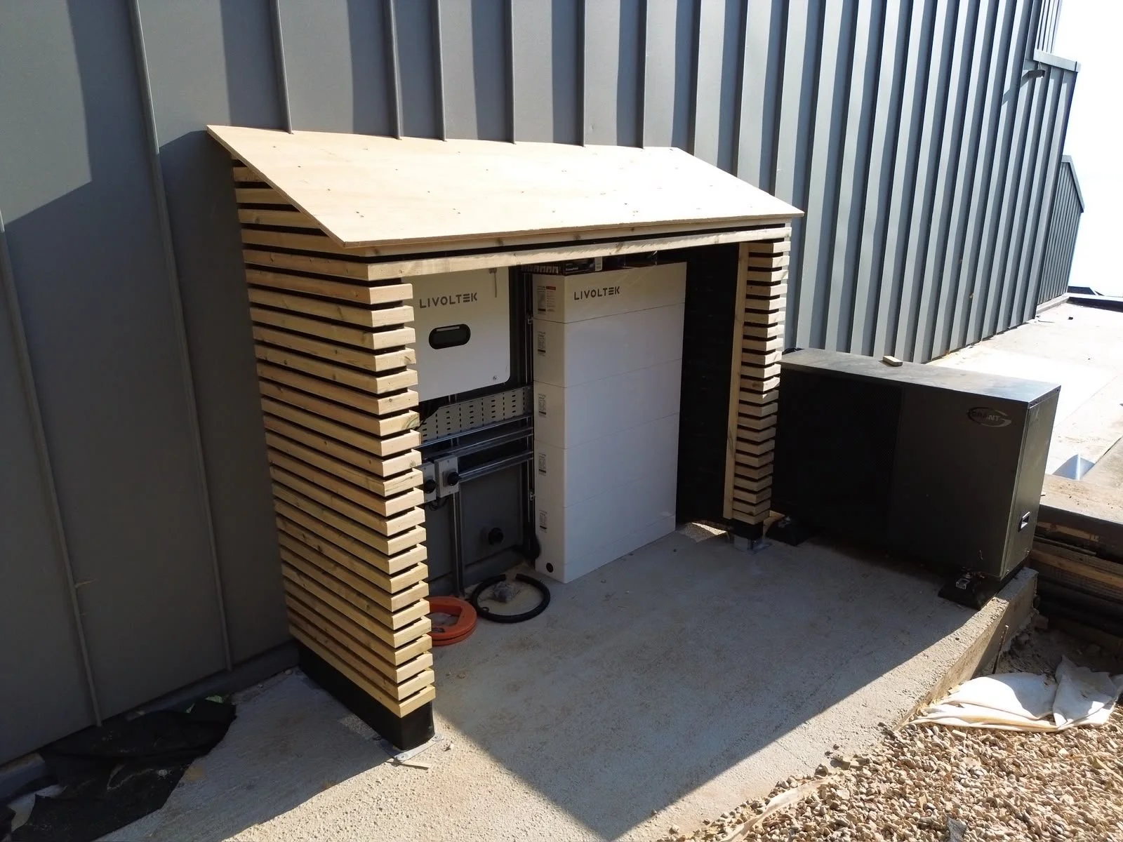 Outdoor electrical installation enclosed in a wooden slat structure with electrical equipment inside, next to a metal building wall.