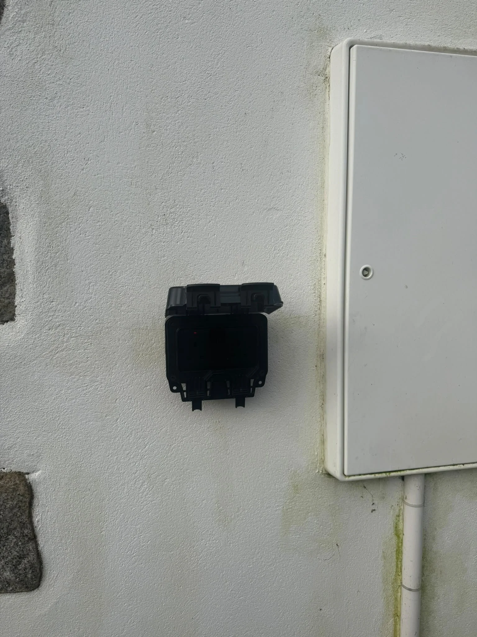 A white electrical box mounted on a textured white wall with a white pipe beneath it and a small black device mounted next to the box.