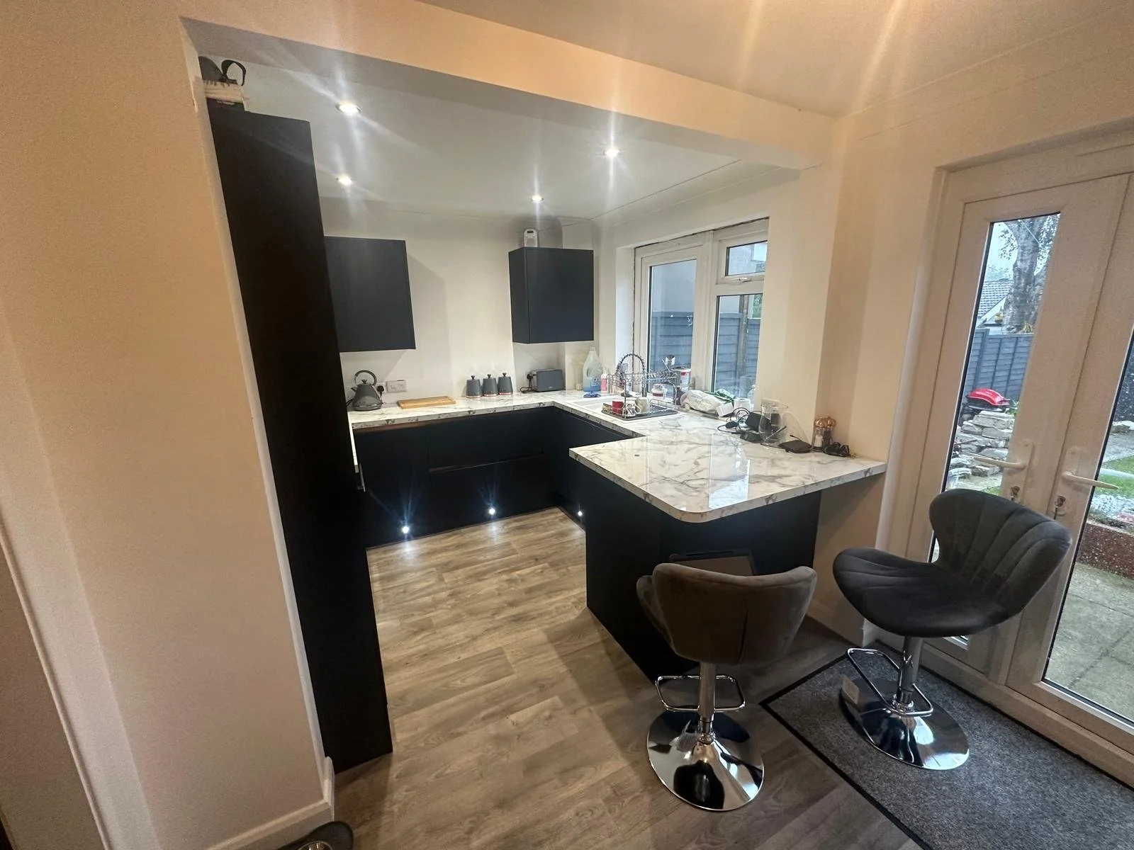 Modern kitchen with black cabinets, marble countertops, and wooden floor. Small dining area with two nearby bar stools. Large glass door leading outside. Various kitchen appliances and items on the counter.