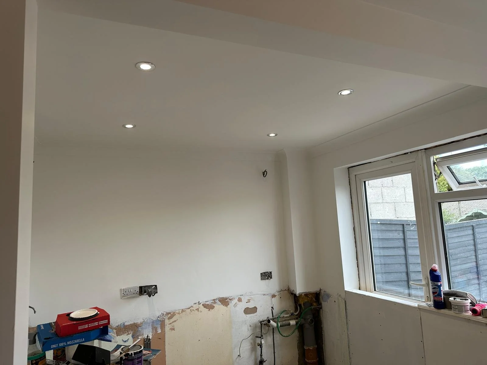Interior of a room under renovation, with a white ceiling and walls, a window with a view outside, and tools and materials scattered around.