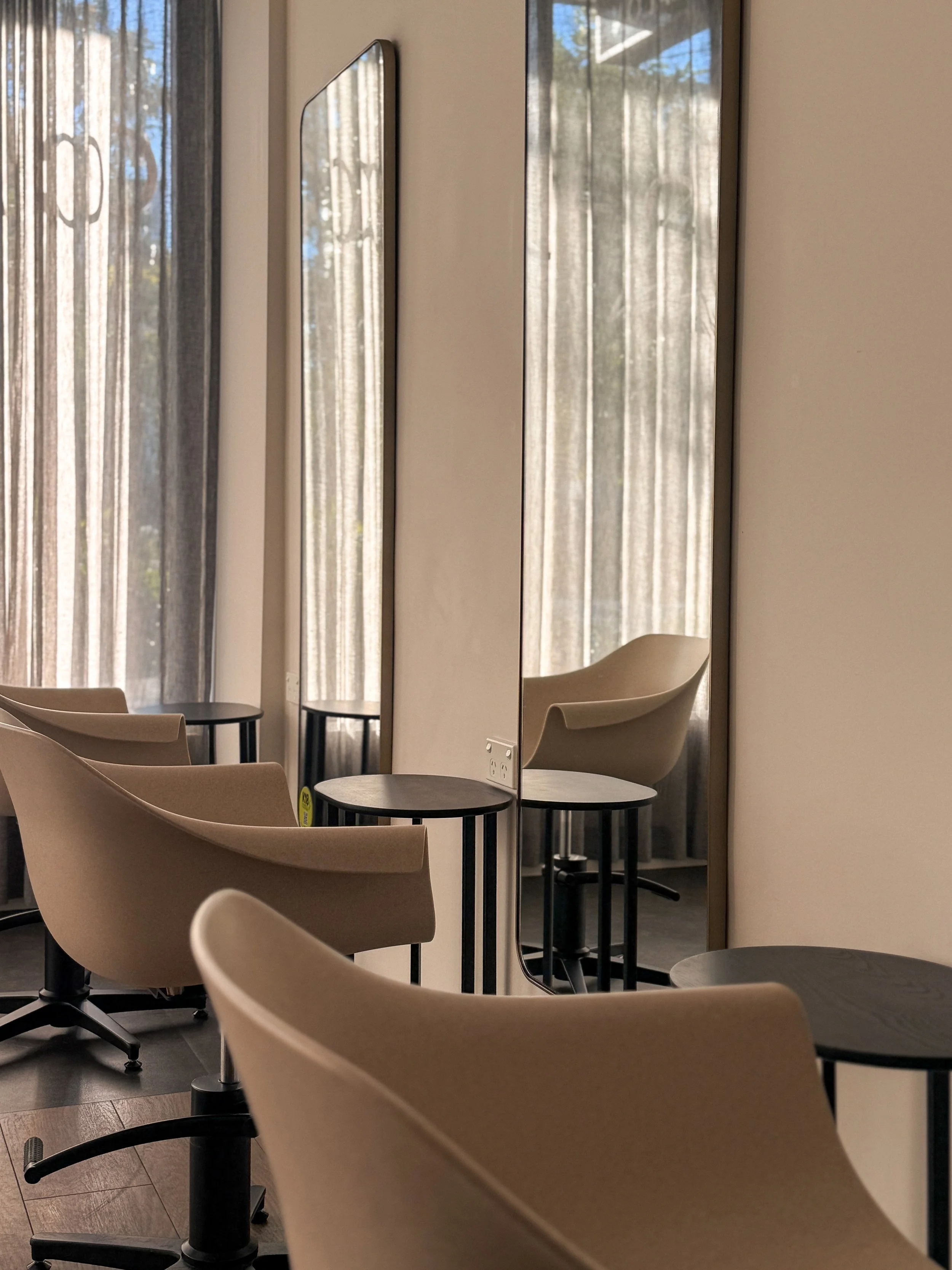 Interior of a waiting area with beige chairs, a small round table, rectangular mirrors on the wall, and large windows with sheer curtains allowing natural light into the space.